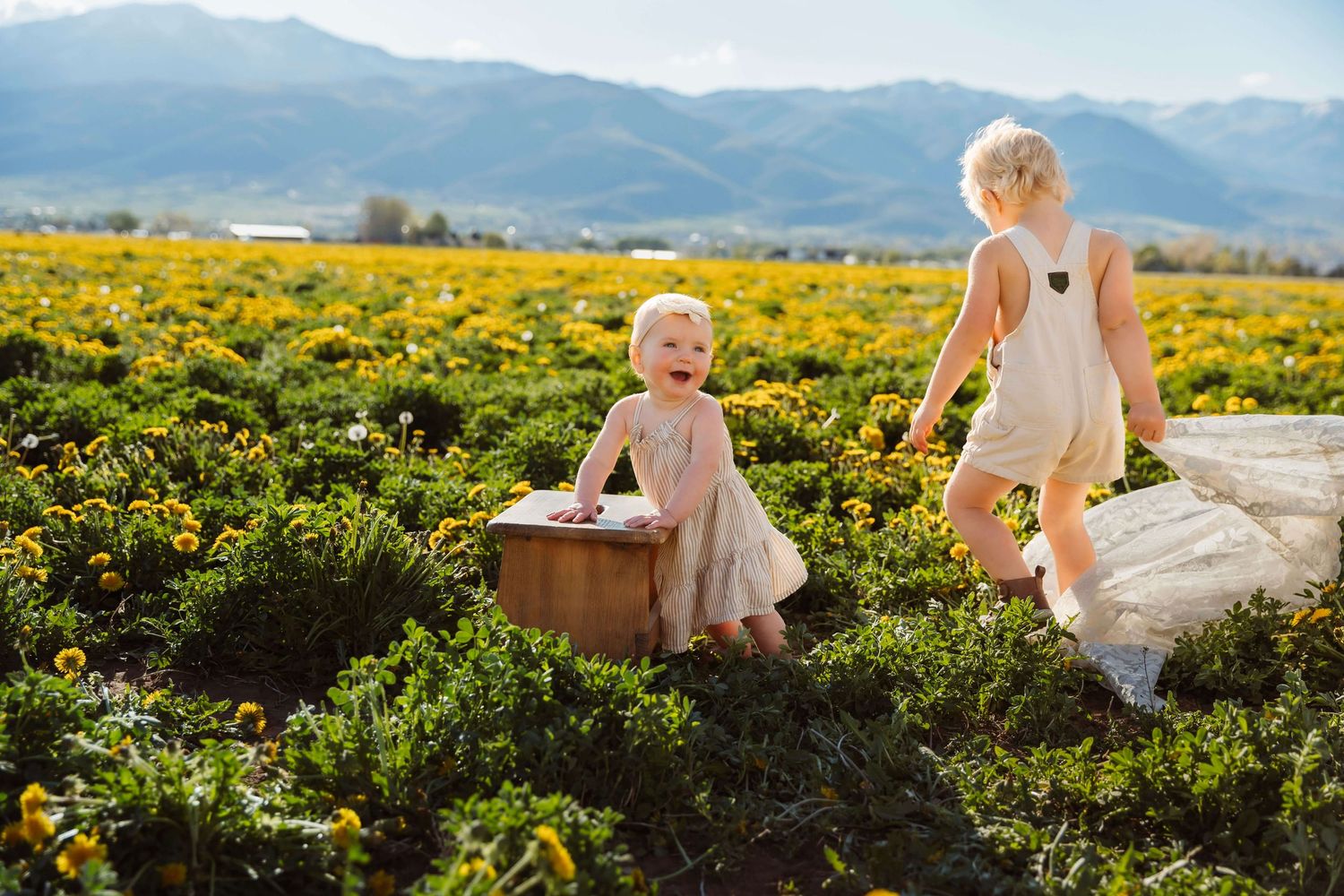 Two children in white outfits exploring a field of yellow wildflowers with mountains in the background.