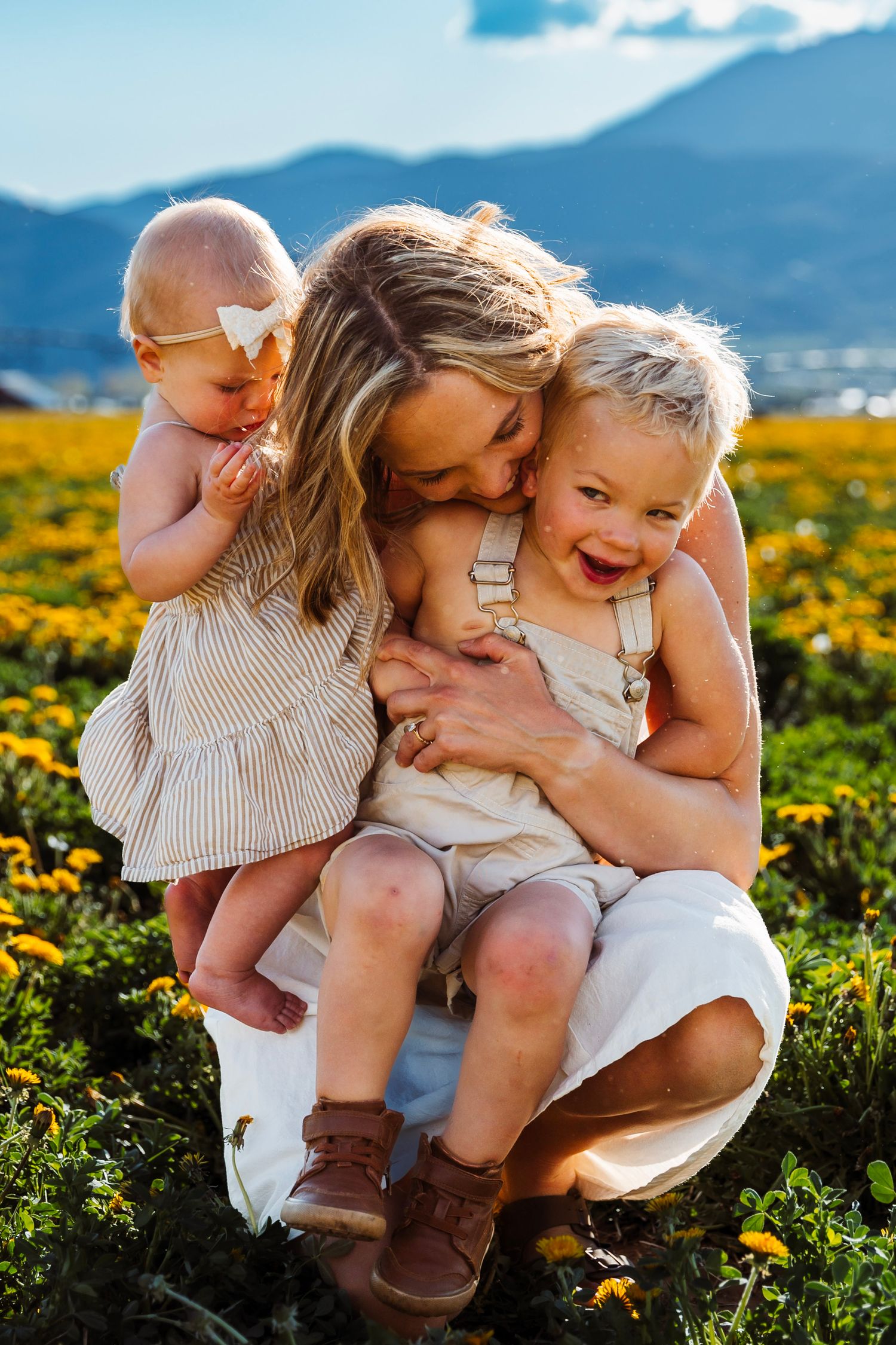 A mother and her children sharing tender moments together in a field of yellow flowers with mountains in background.