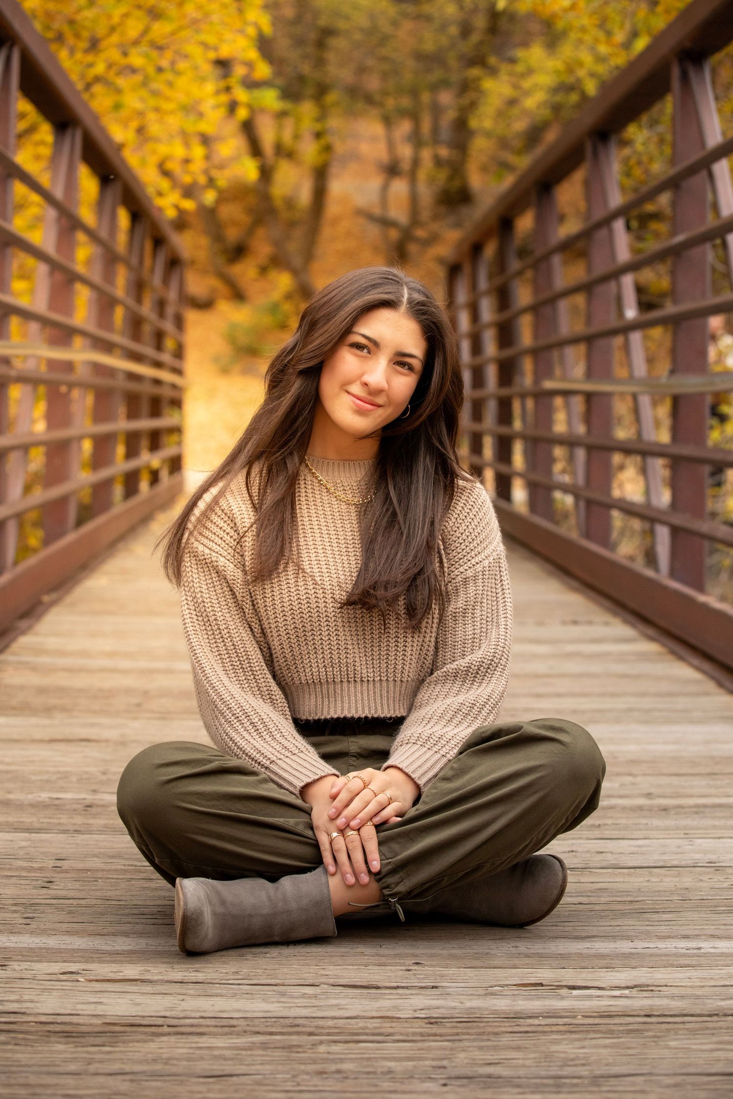 Woman in beige sweater and olive pants poses on wooden bridge with golden autumn foliage in background.