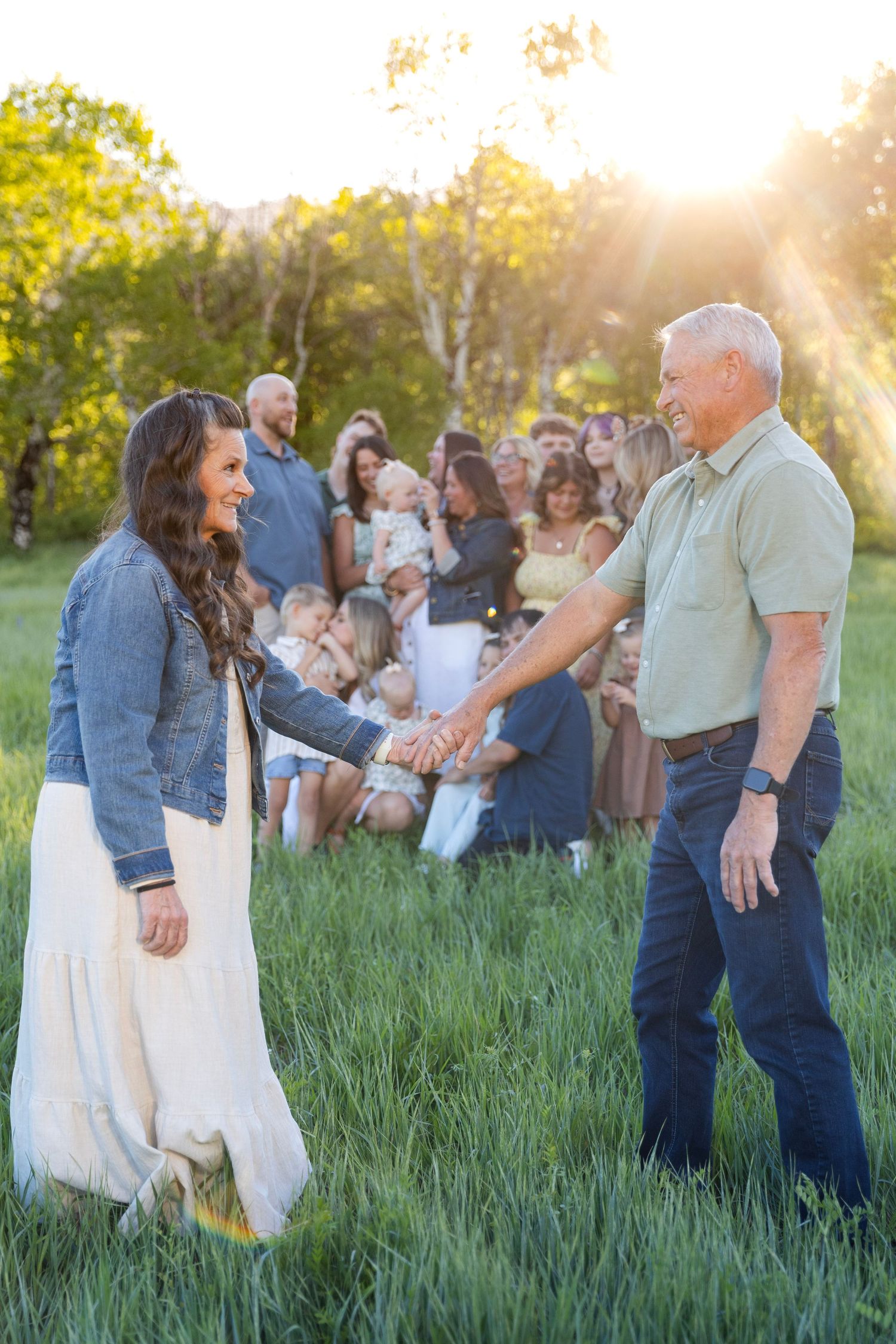 A group of people gather in a grassy field at sunset as two individuals stand together in warm evening light.