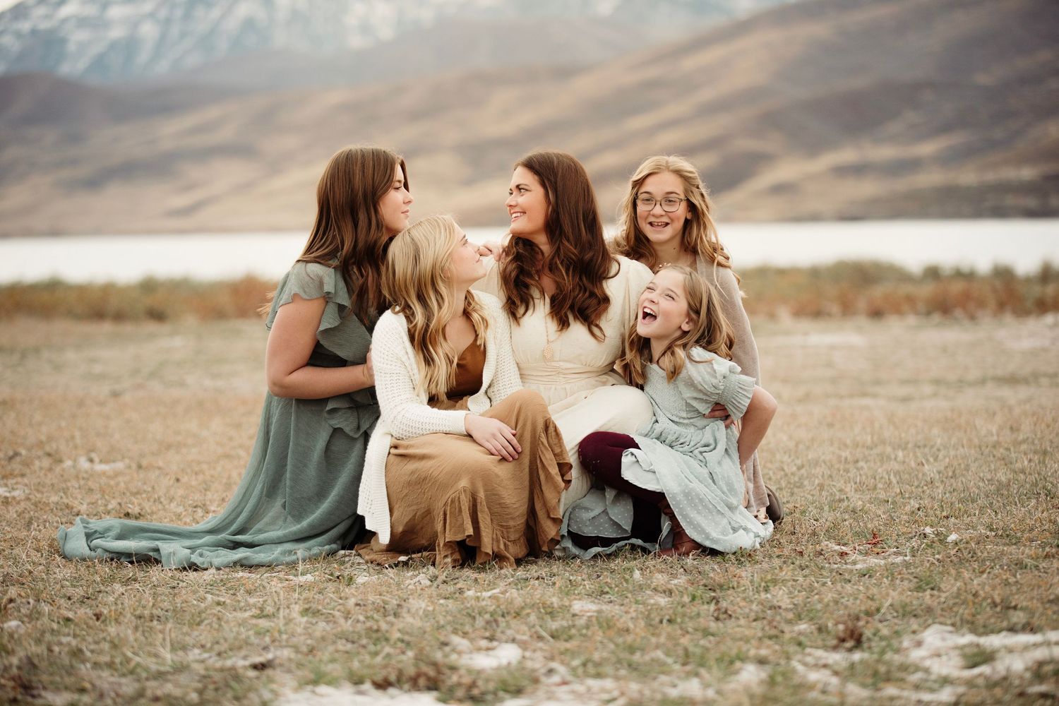 A mother and her daughters sitting together on the ground in a natural outdoor setting with mountains in the backdrop at sunset.