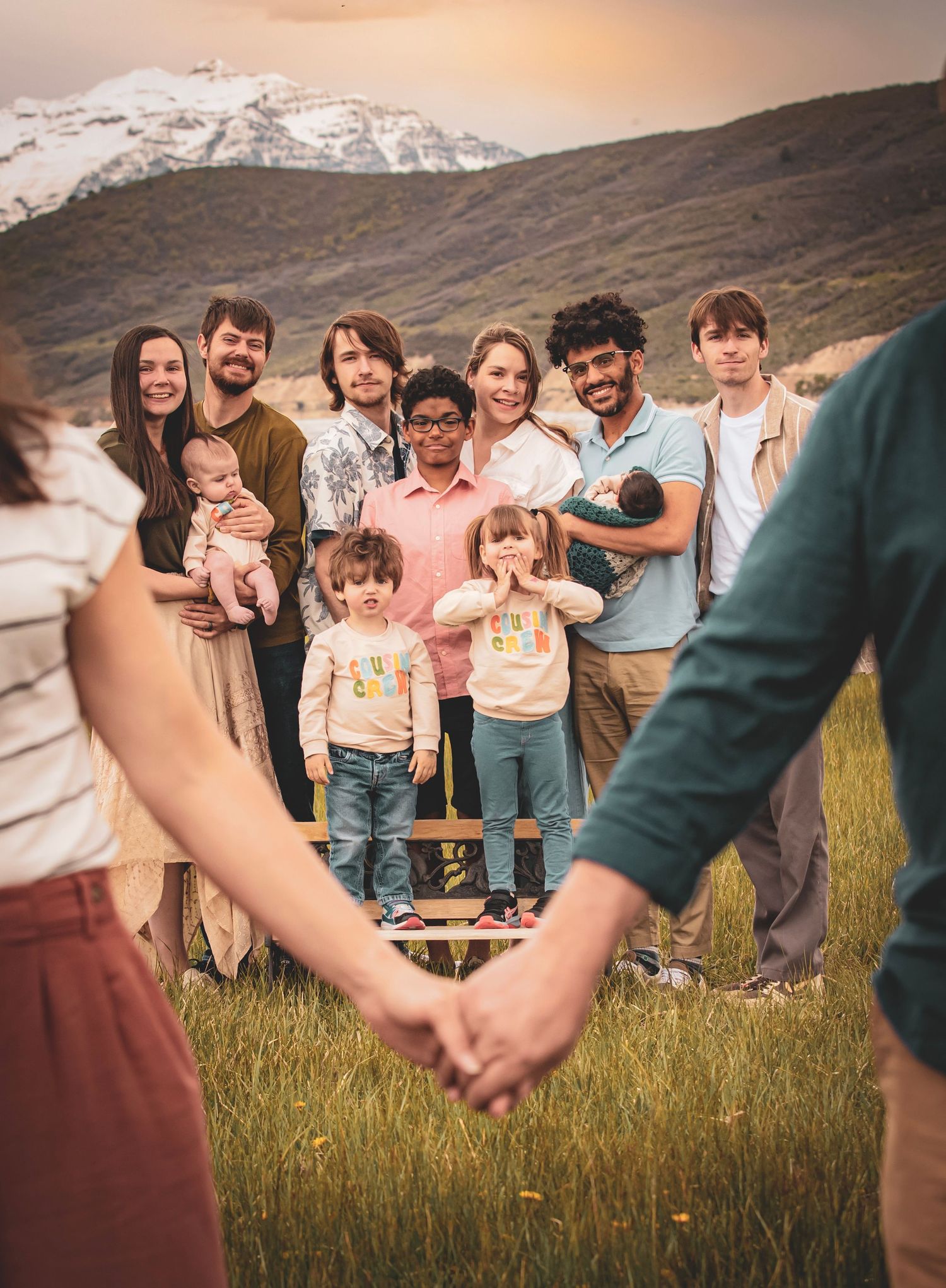 A candid outdoor family photo showing a group posing on grassy terrain against snowy mountains at sunset.