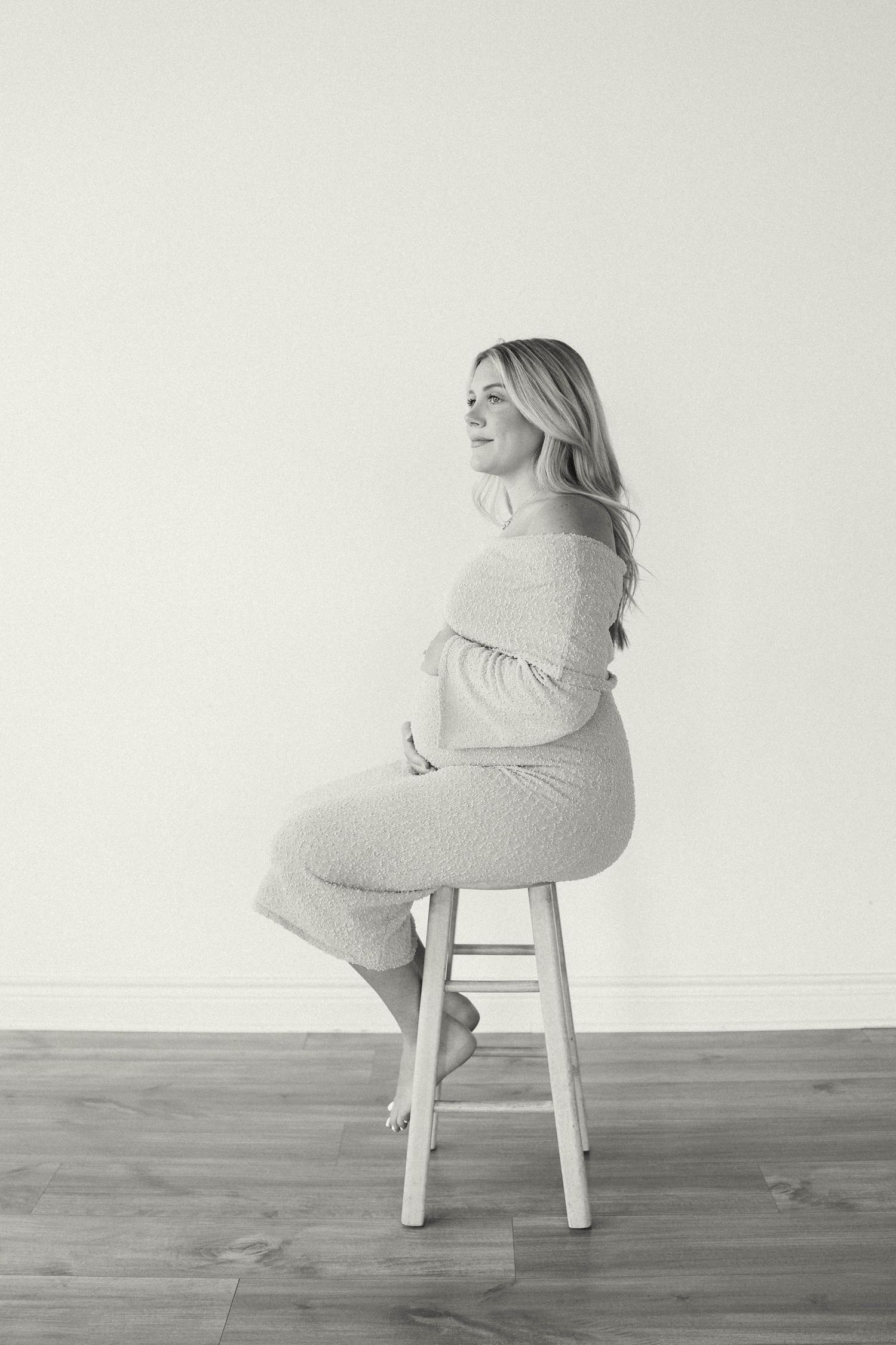 Black and white photo of a pregnant woman sitting on a stool, cradling her baby bump in a cozy knit outfit.