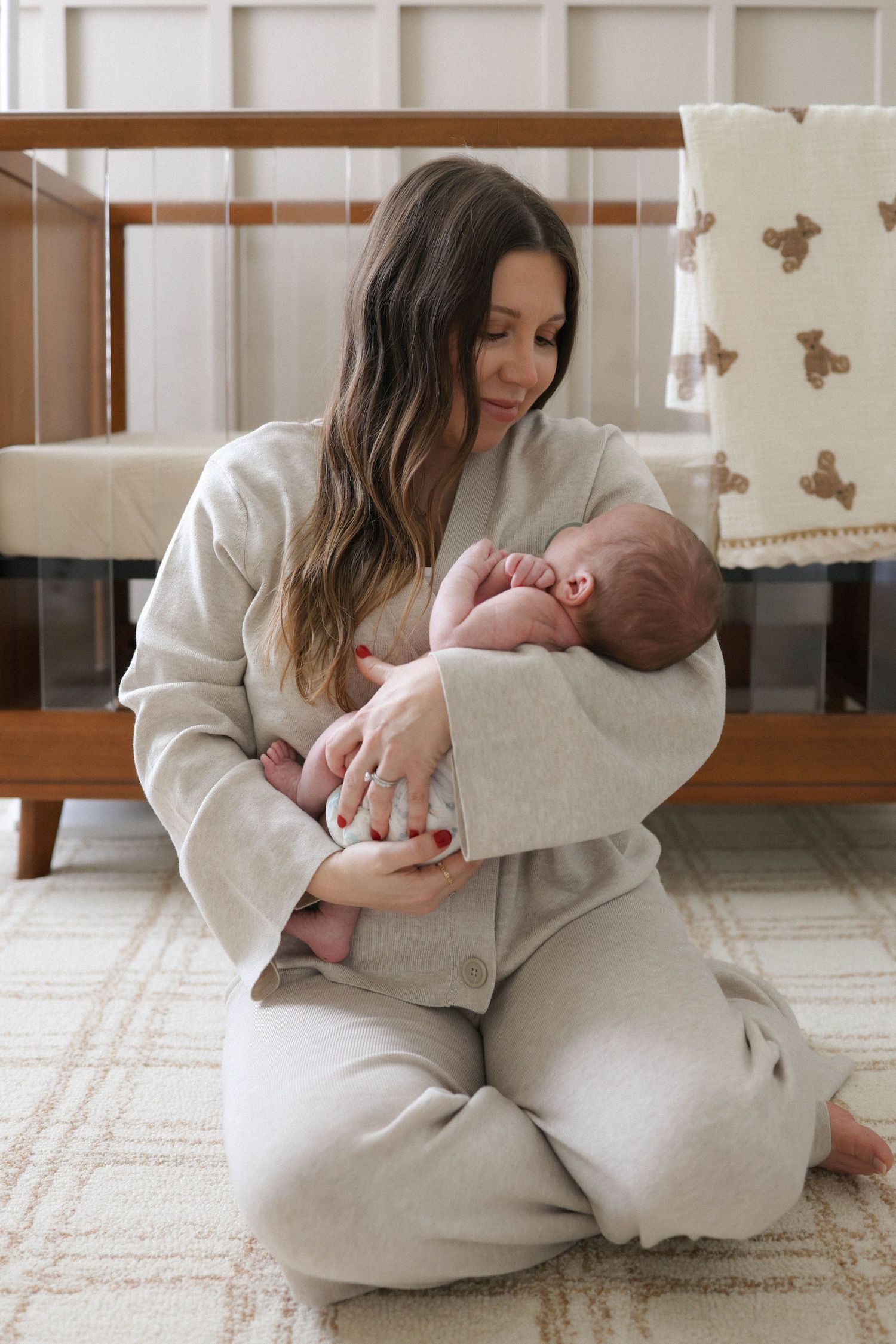 A mother in a gray loungewear set sits on the floor cradling her newborn baby in a nursery.