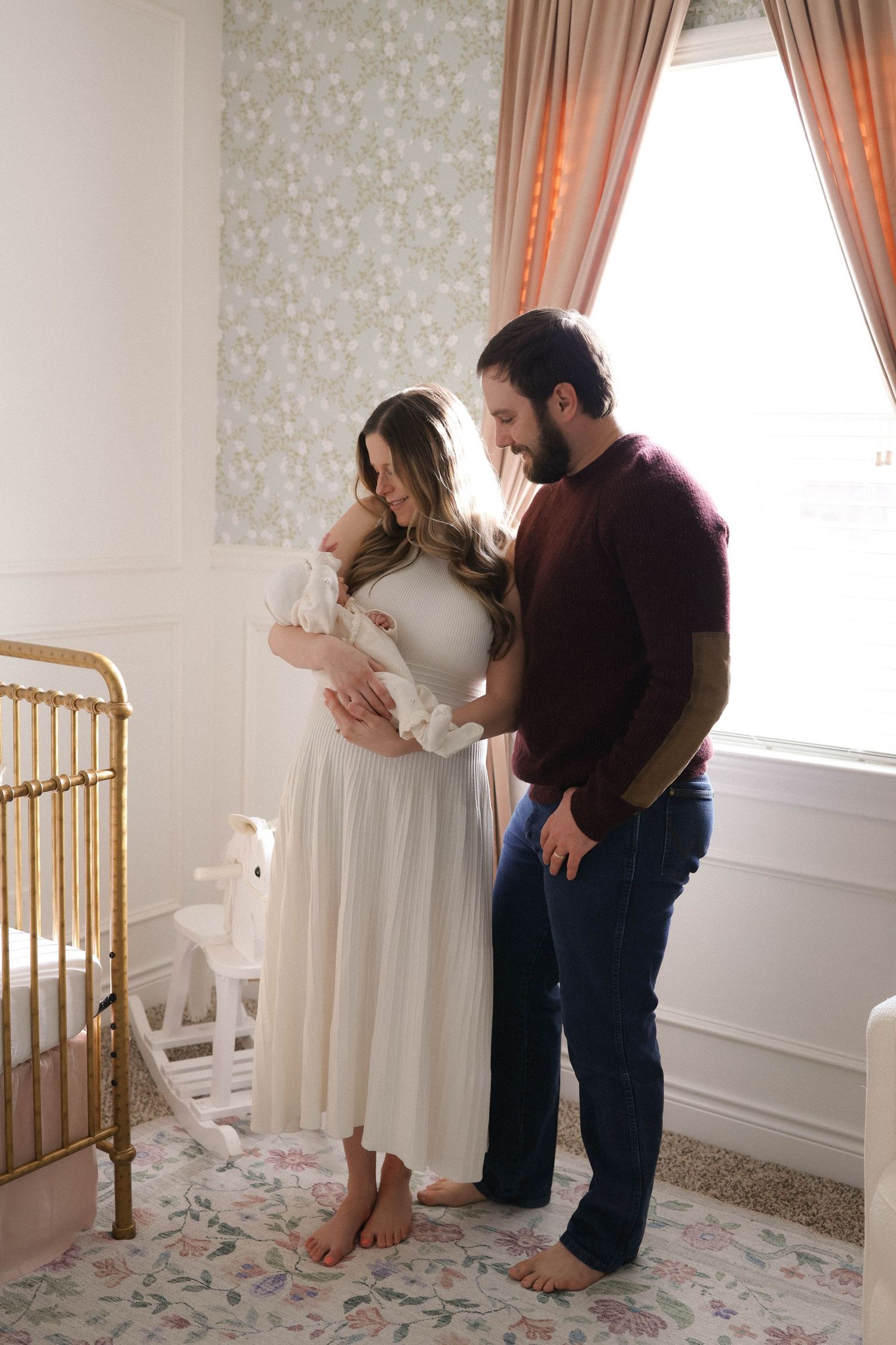 A couple admires their newborn baby in a beautifully decorated nursery with floral wallpaper and a gold crib.