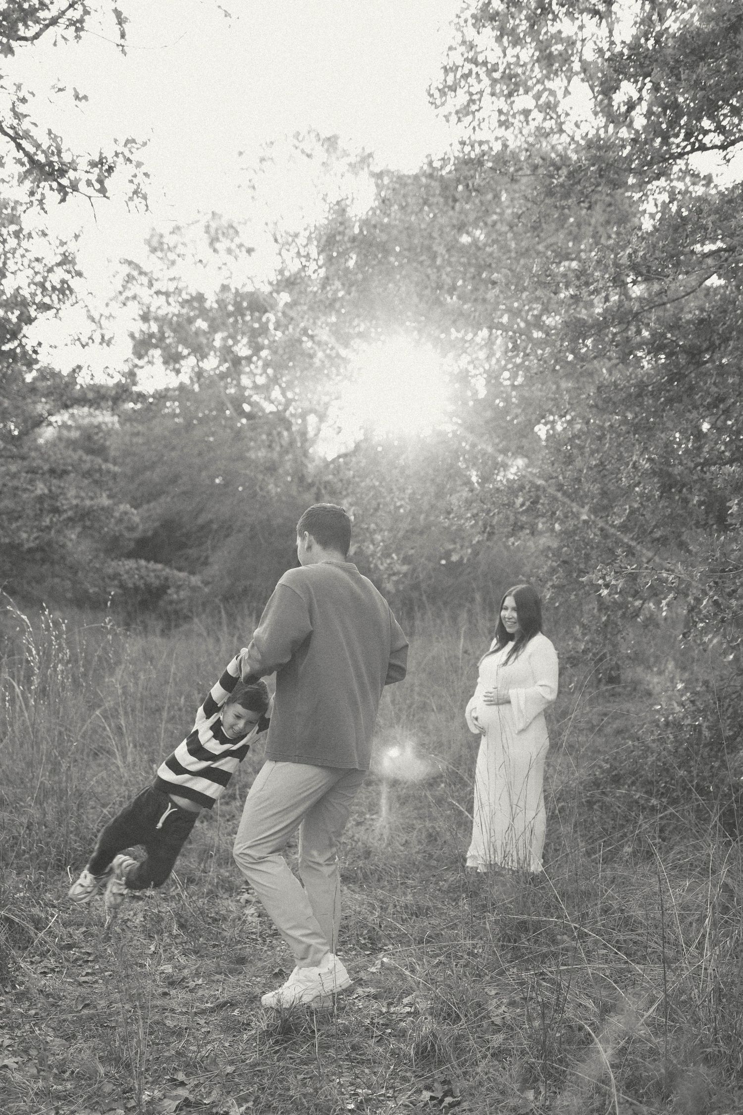 Black and white maternity photo of pregnant woman in lace dress with partner and child in sunlit forest clearing.