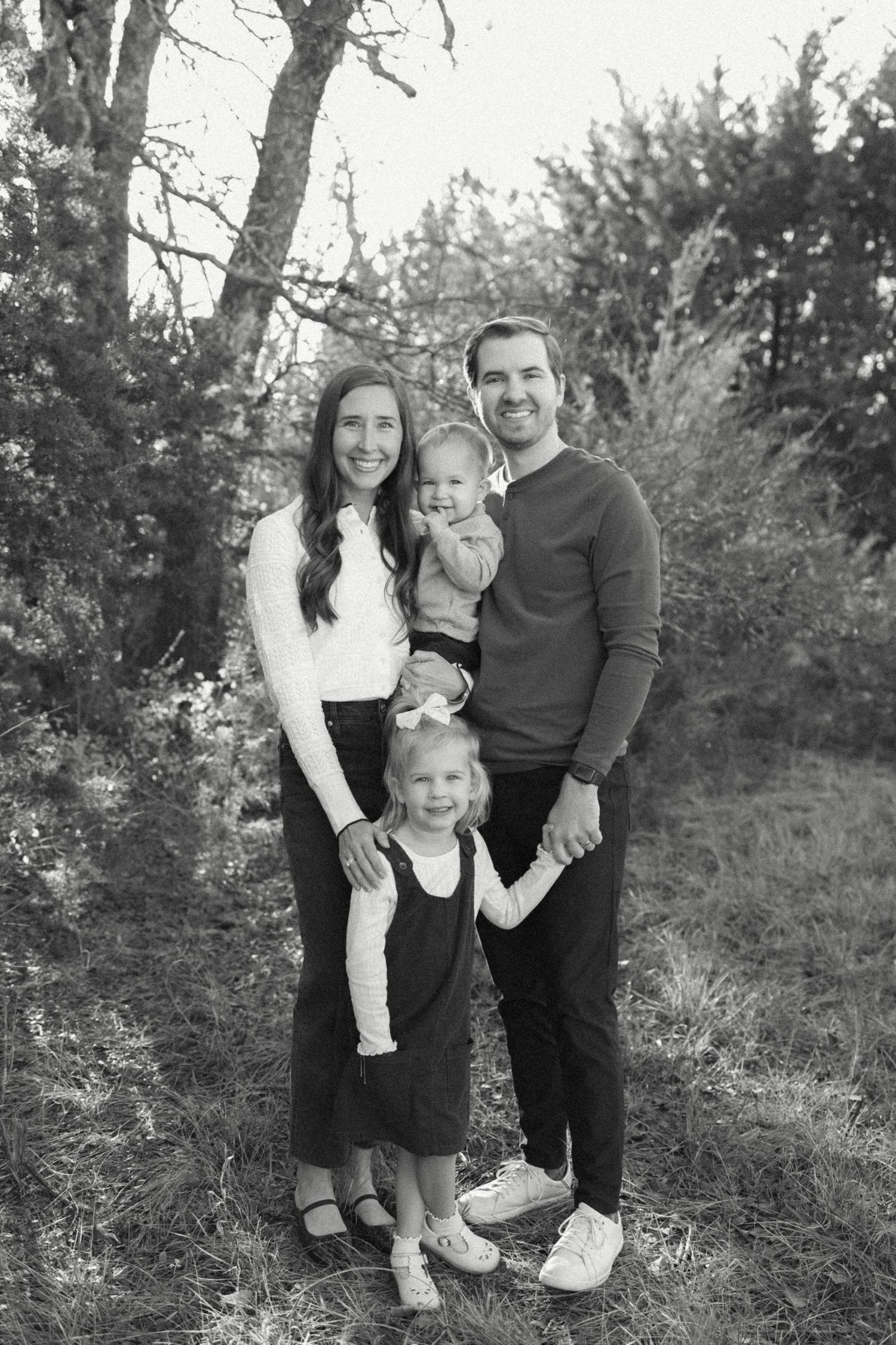 Black and white family portrait of parents with two young children posing outdoors among trees.