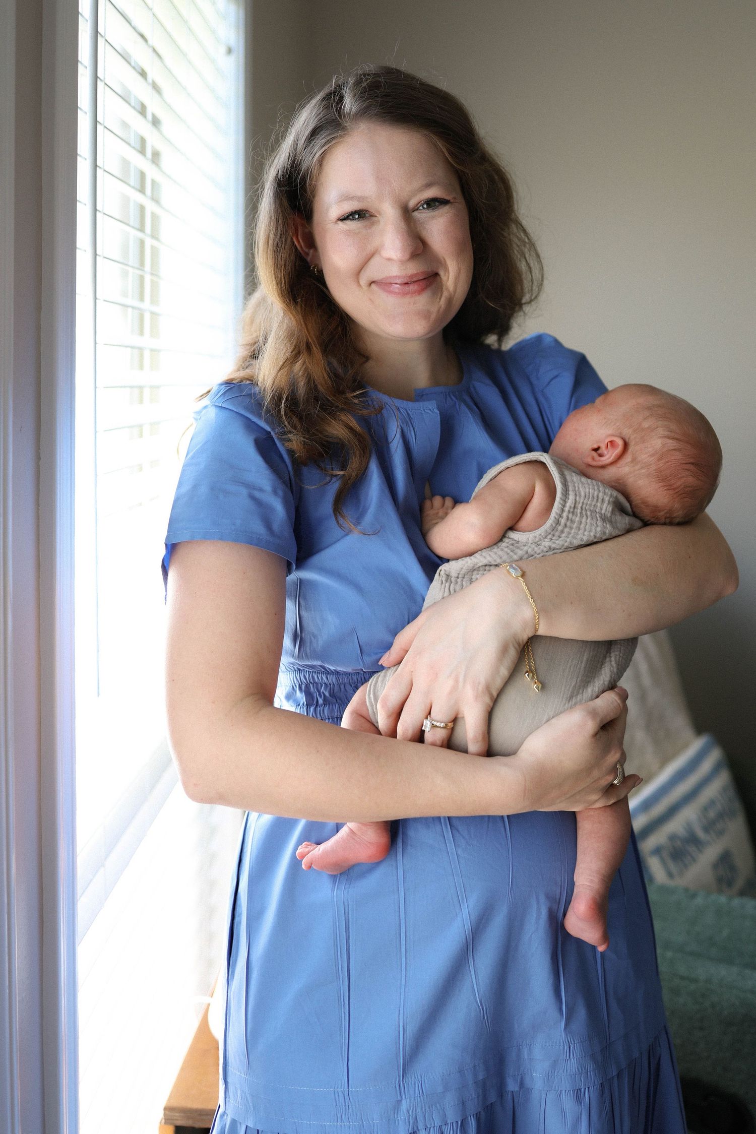 Smiling woman in blue dress holding a newborn baby near a window indoors.