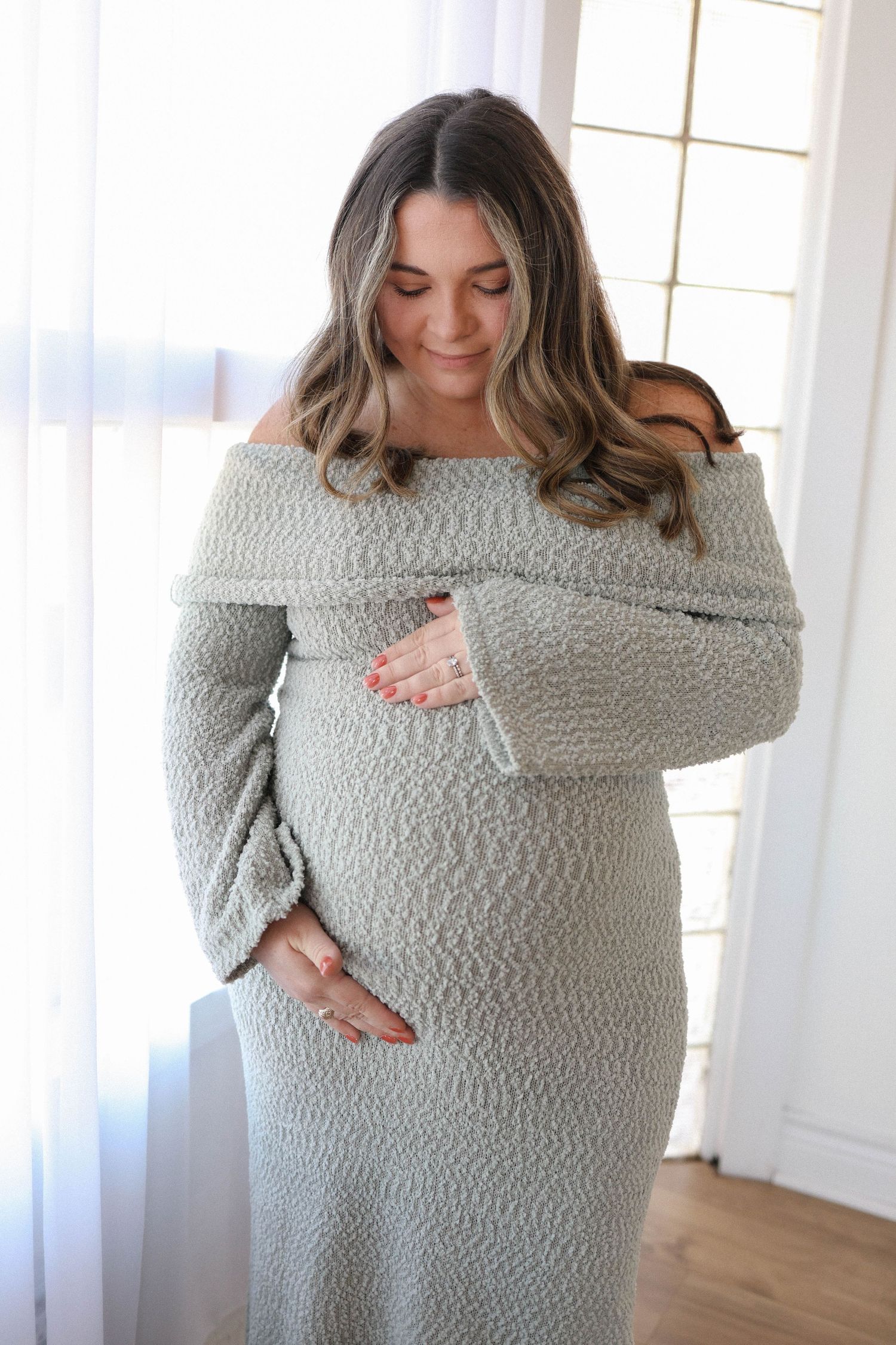 Pregnant woman in off-shoulder sage green knit dress cradling baby bump, standing by window.