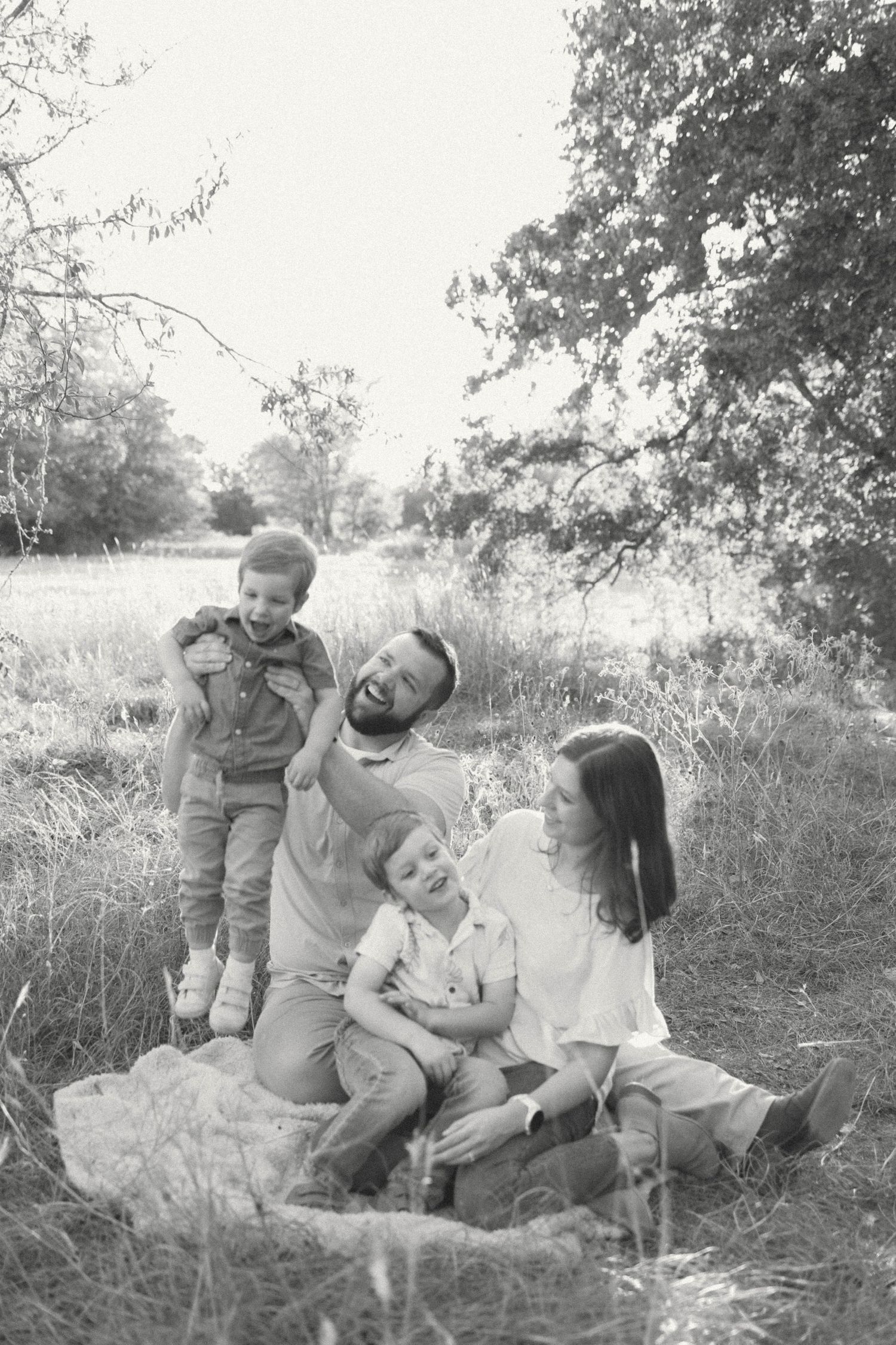 Black and white photo of a happy family with two young boys playing outdoors in a grassy field near trees.
