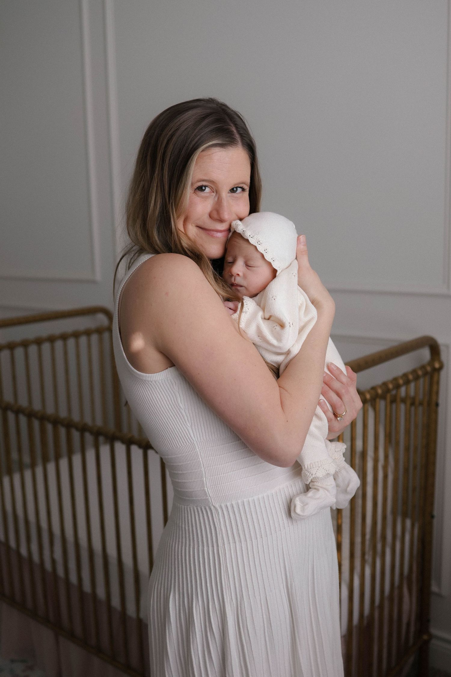 A mother in a white dress tenderly holds her sleeping newborn baby in a white bonnet near a gold crib.