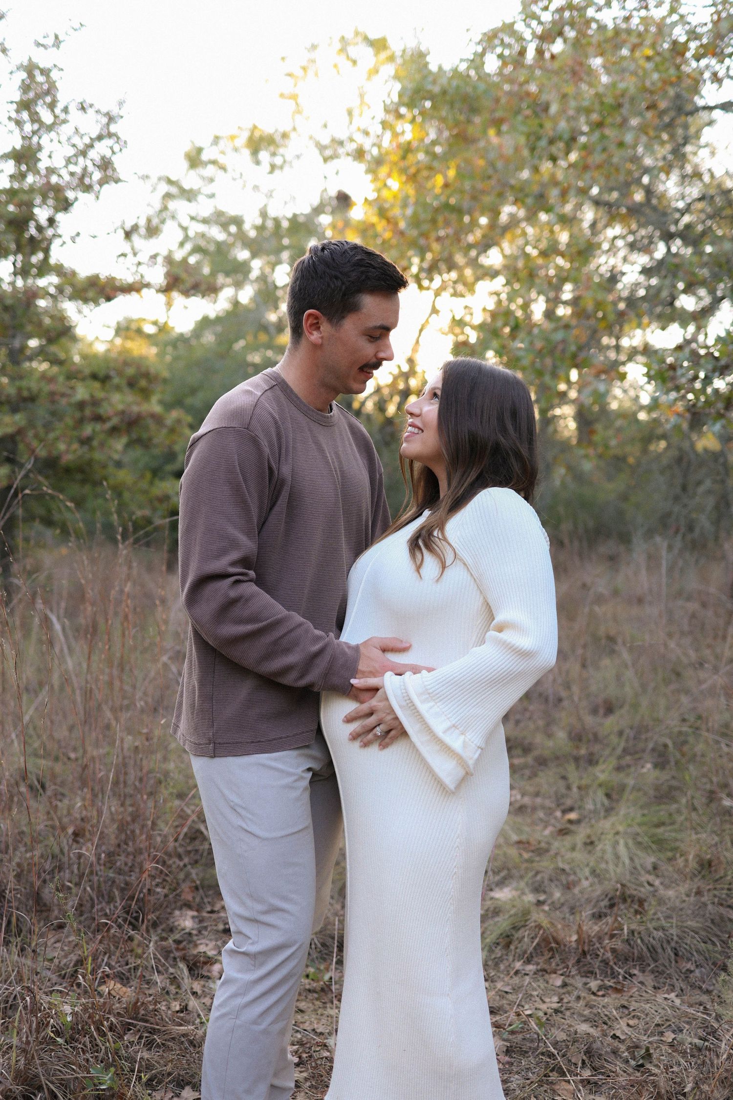 Couple sharing a tender moment outdoors, pregnant woman in white dress, man in brown sweater, golden hour forest backdrop.