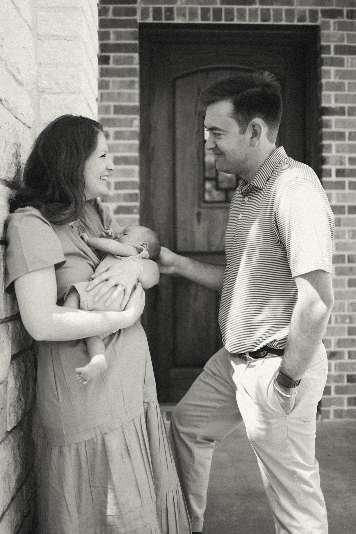 Black and white photo of a couple smiling at each other while the woman holds a newborn baby outside their home.