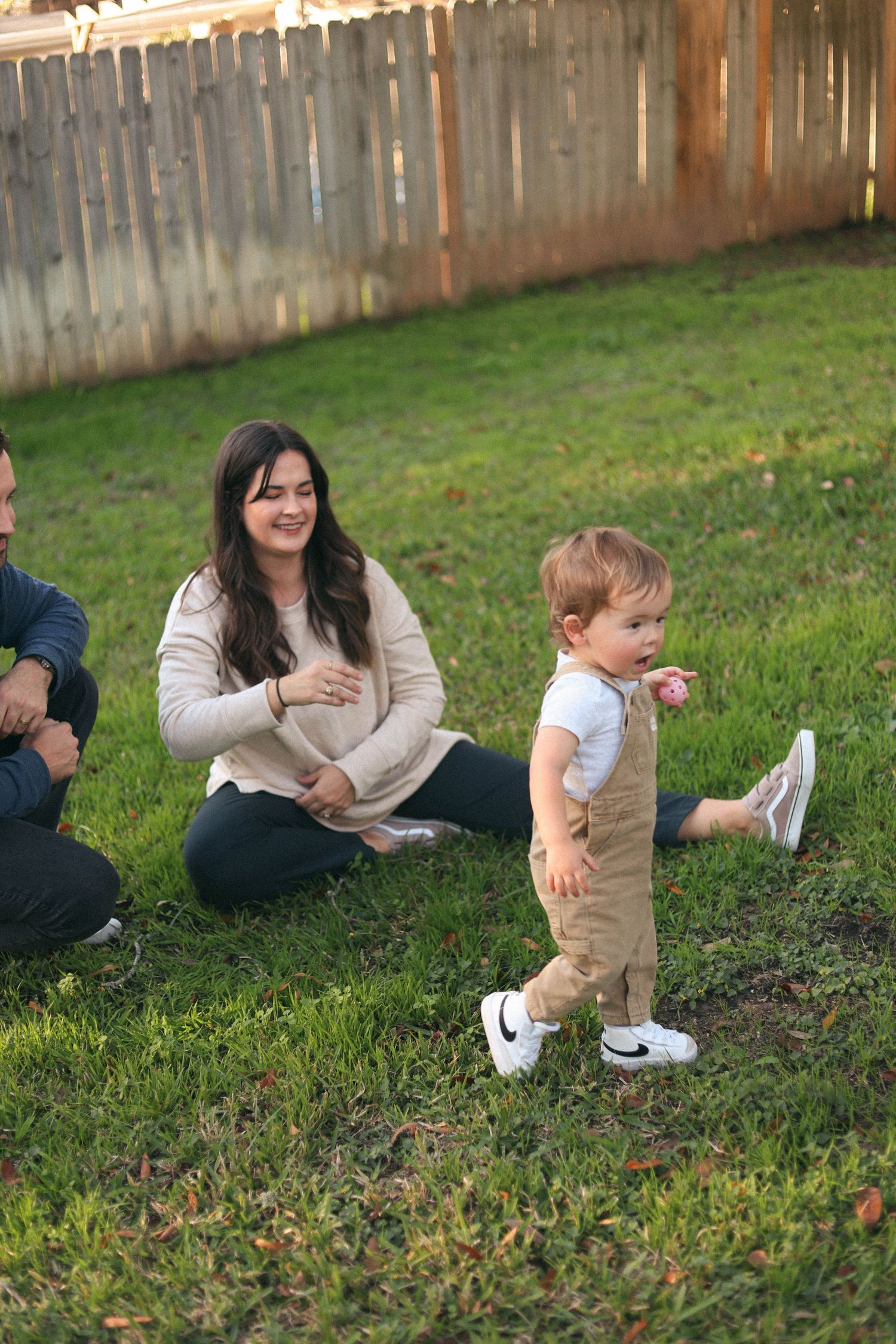 Toddler walking in backyard while parents sit and watch.