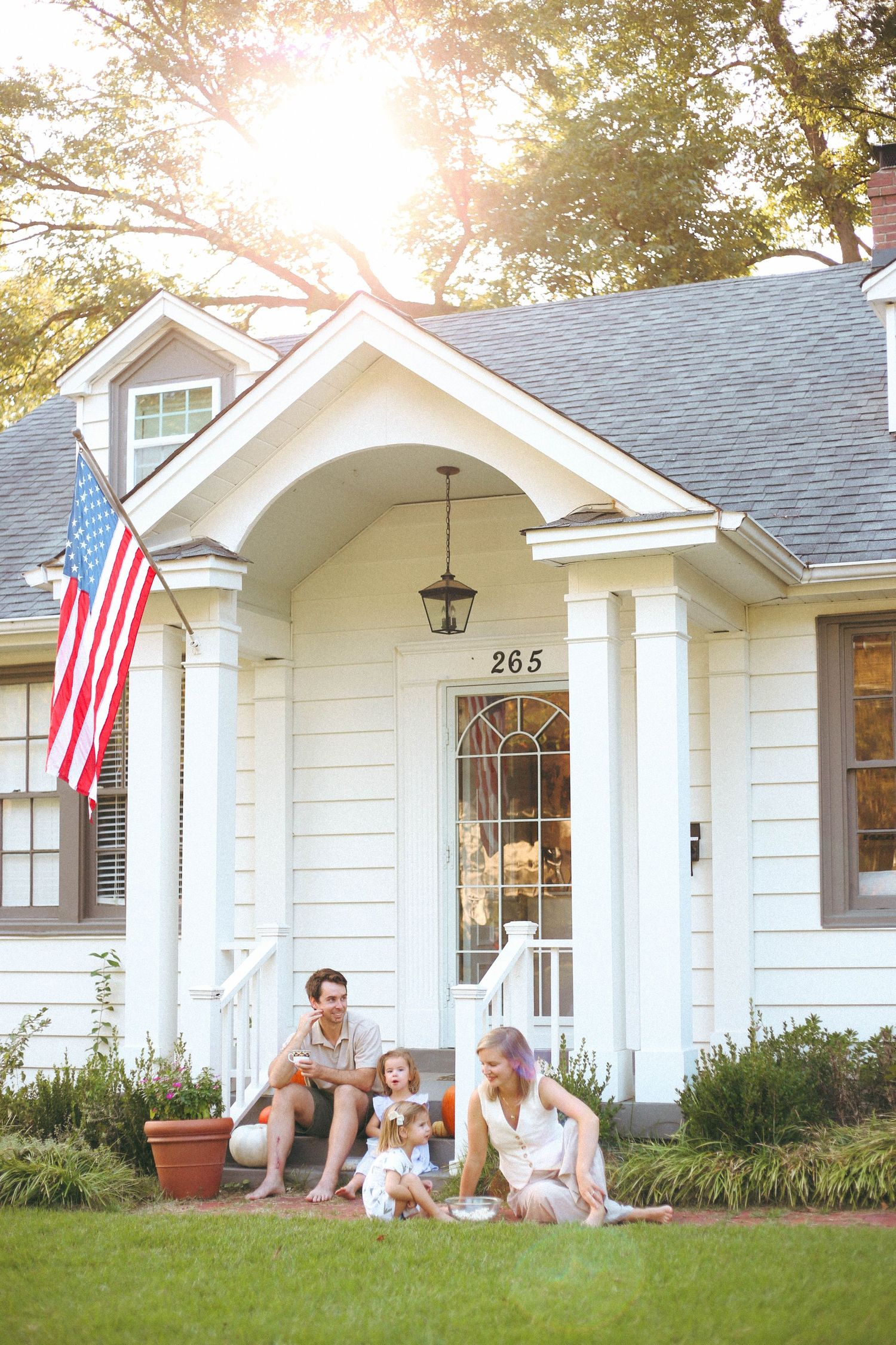 A family relaxes on their front lawn in front of a classic white house with American flag.