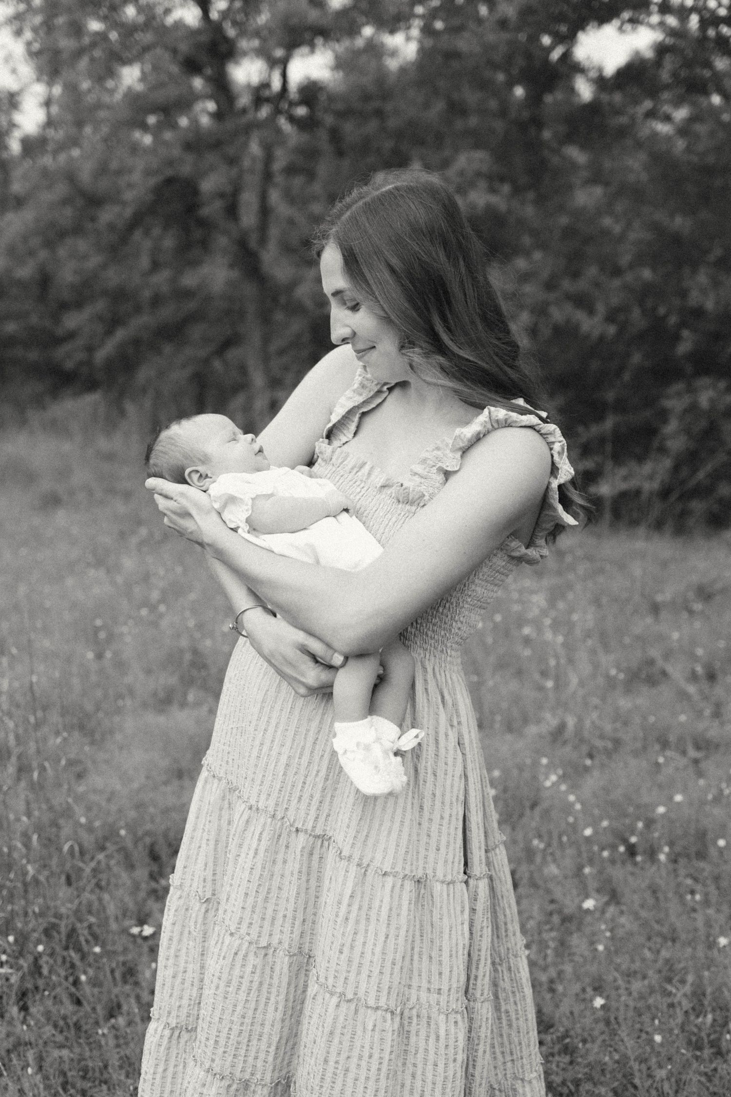 Black and white photo of a smiling mother in a maxi dress cradling her newborn baby outdoors in a lush green field.