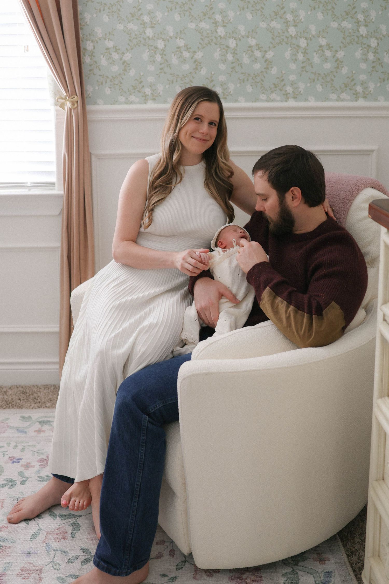 A couple sits in a white nursery chair, lovingly gazing at their newborn baby in a floral-wallpapered room.