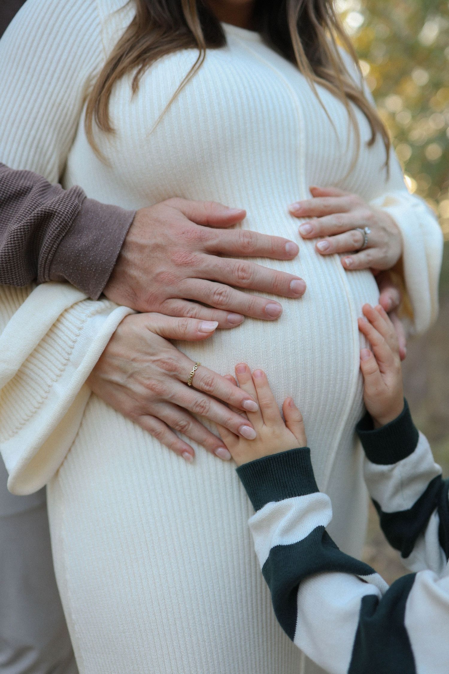 Pregnant woman in white ribbed dress with multiple hands gently cradling her baby bump outdoors.