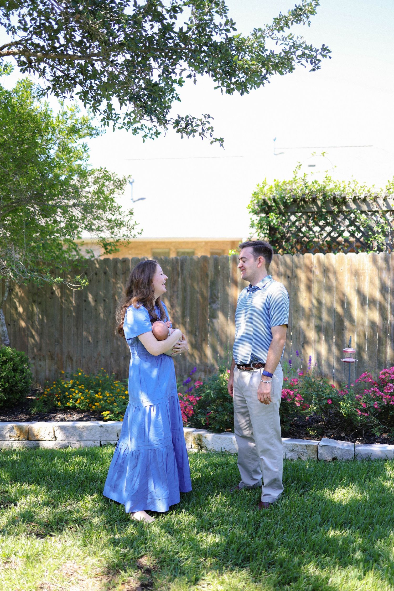 A couple stands in a sunny backyard, the woman in a blue maxi dress holding a newborn baby, smiling at each other.