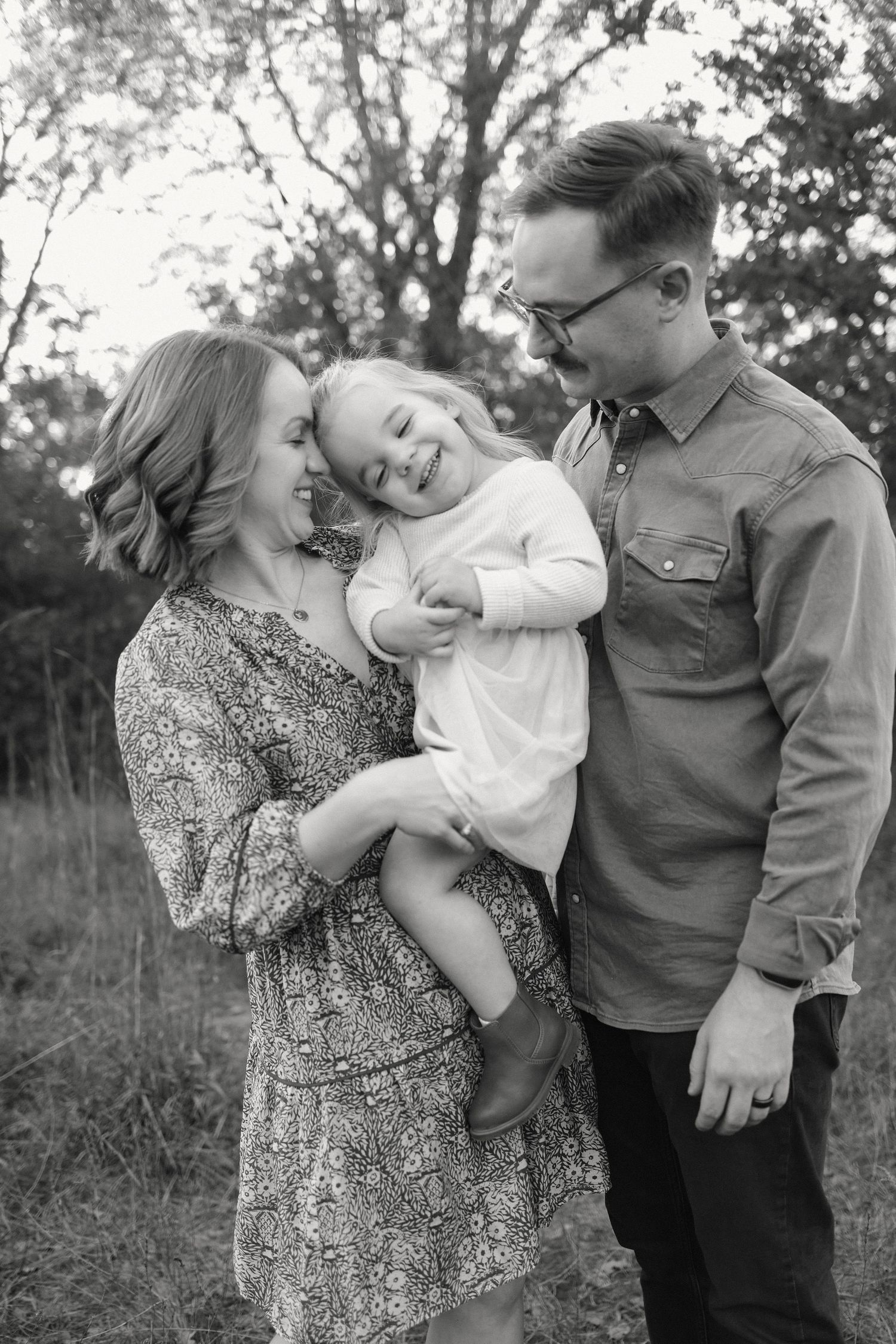 Black and white family portrait of parents smiling at laughing toddler girl held in mothers arms outdoors.