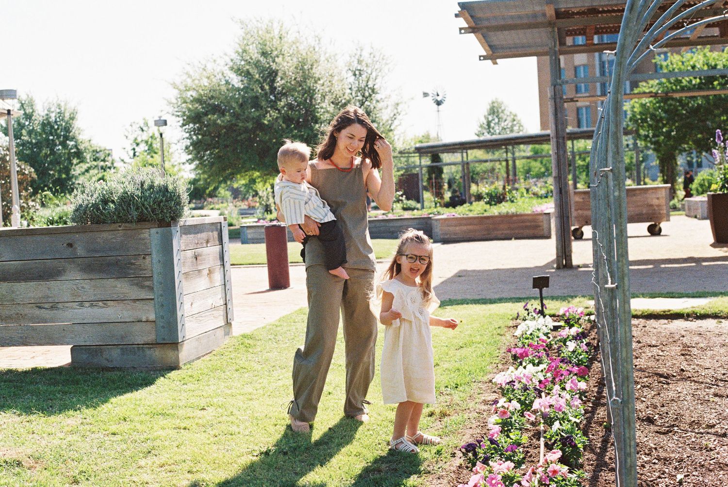 A mother carrying a baby walks with a young girl through a lush garden with pink flowers and wooden planters.