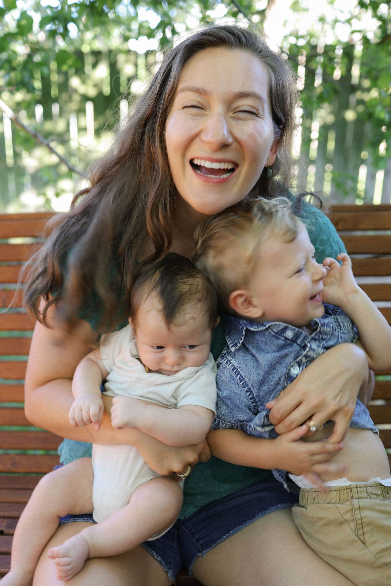 Laughing mother sitting on wooden bench outdoors holding two young babies, enjoying a joyful family moment in a garden.