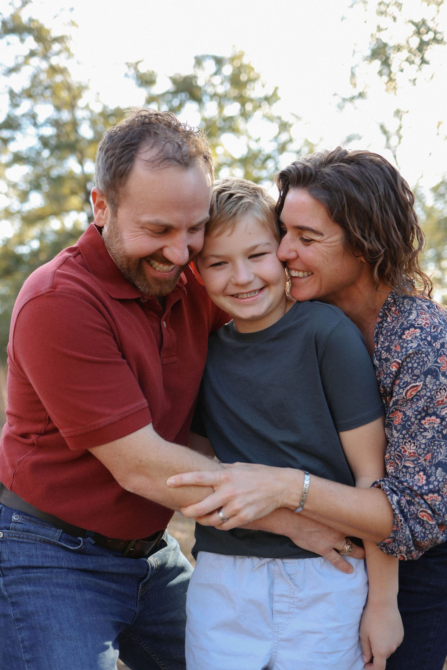 Happy family portrait with parents hugging their smiling son outdoors in natural light.