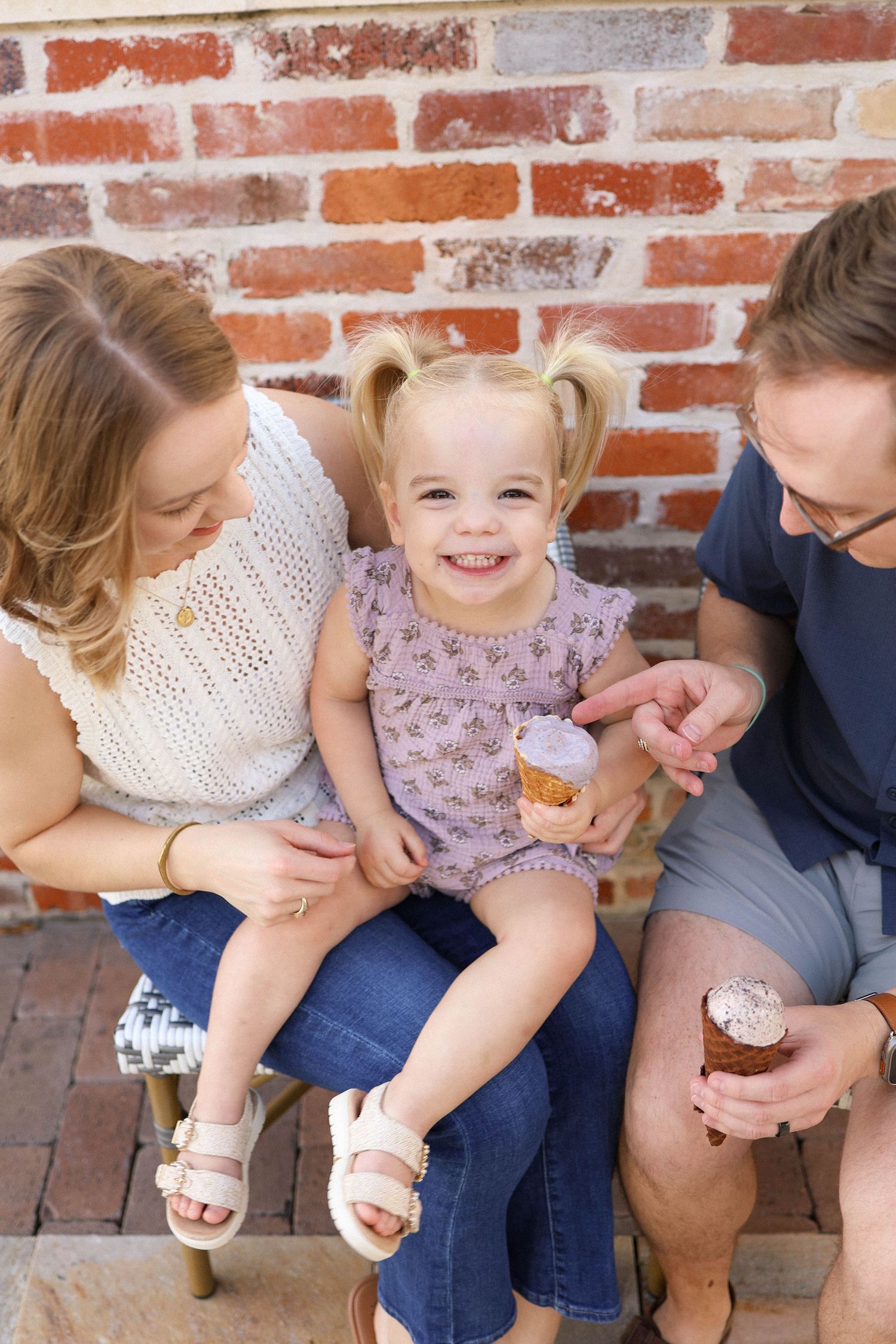 A happy toddler girl with pigtails enjoys ice cream with her smiling parents against a rustic brick wall backdrop.