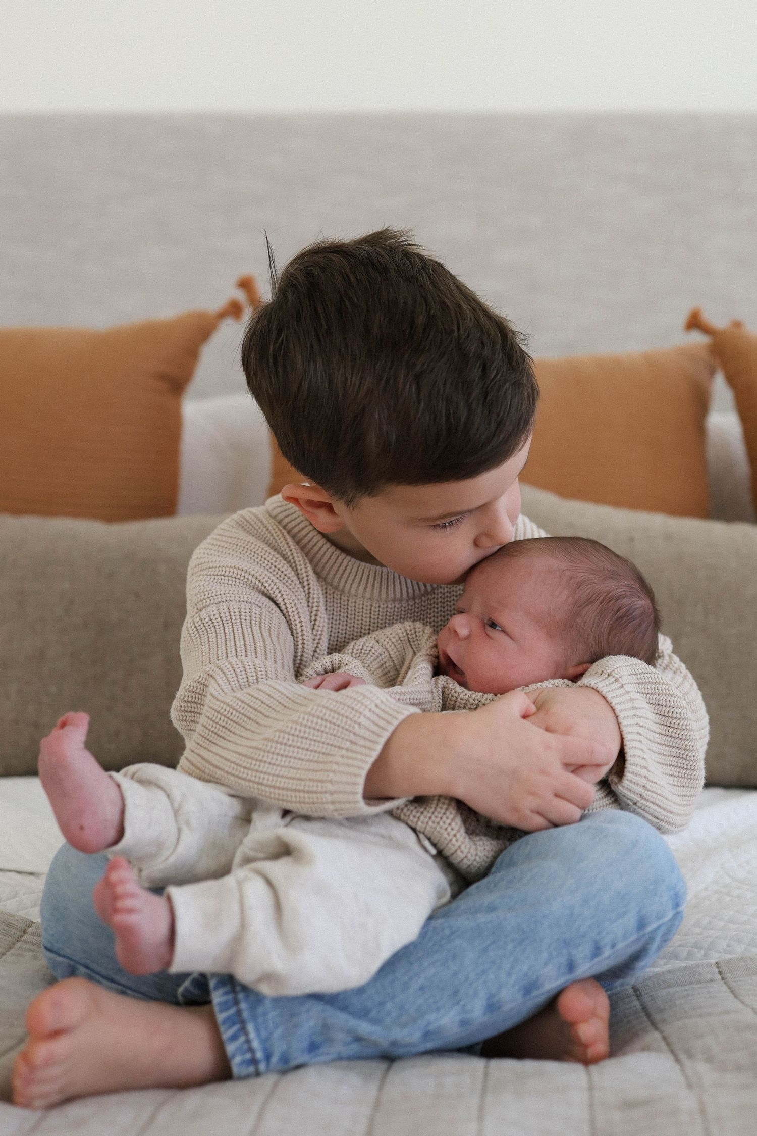 Young boy sitting on a couch gently kissing a newborn baby he holds carefully in his arms.