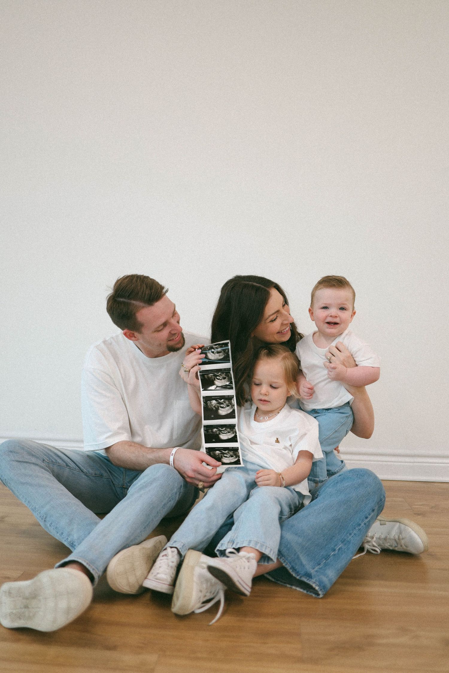 A family sits on wooden floors sharing ultrasound photos while children play.