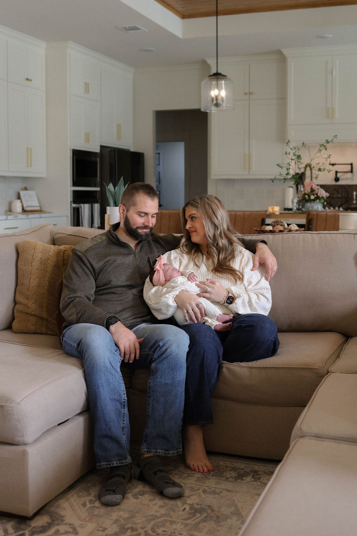 A couple sits on a beige sectional sofa, smiling at each other while holding their newborn baby in a modern home.