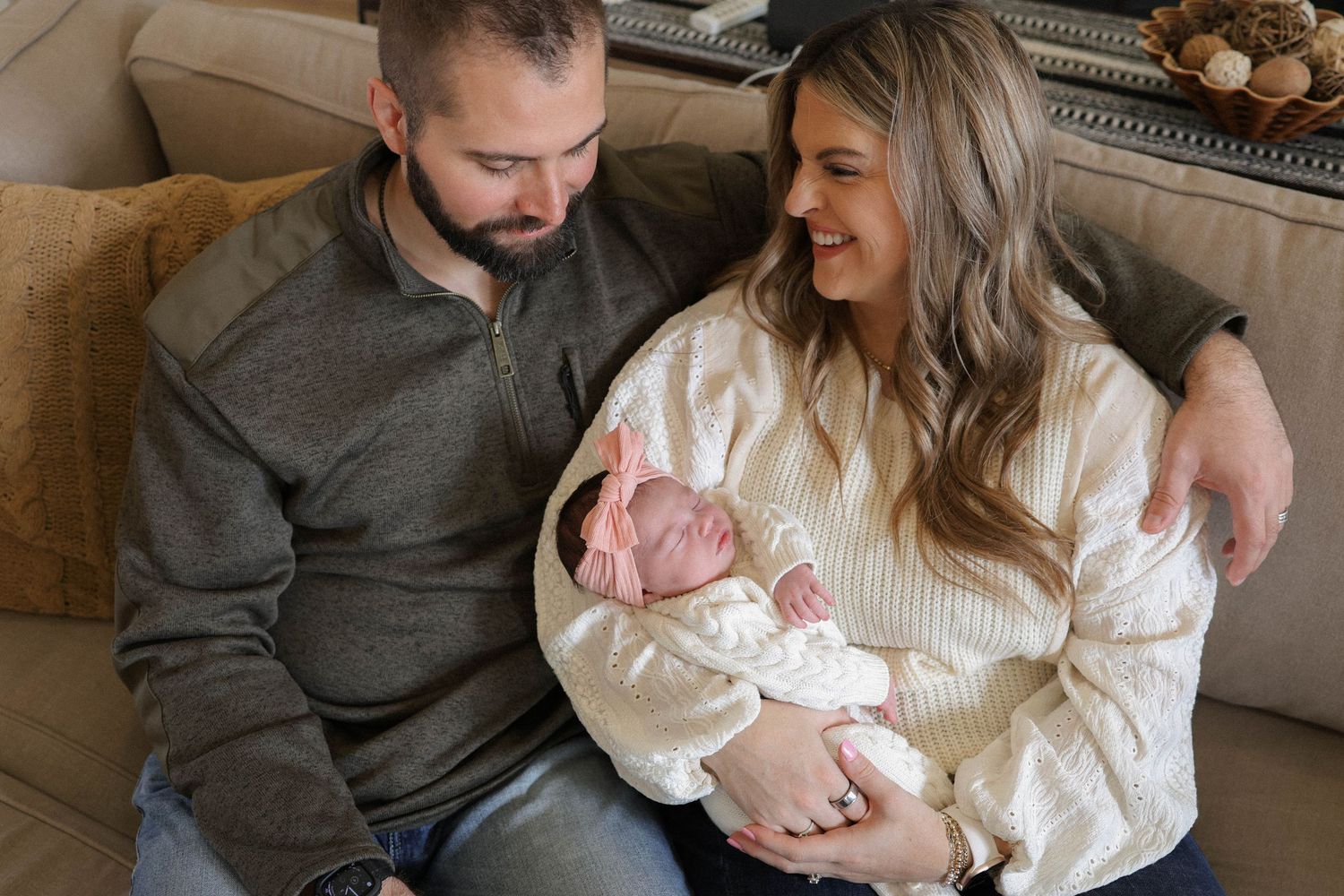 A couple sits on a couch, smiling as they cradle their newborn baby wrapped in white.