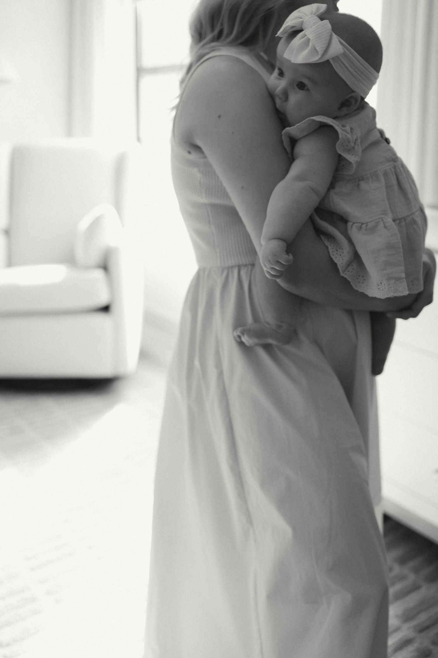 Black and white portraits of mother holding baby in white dress against bright window.