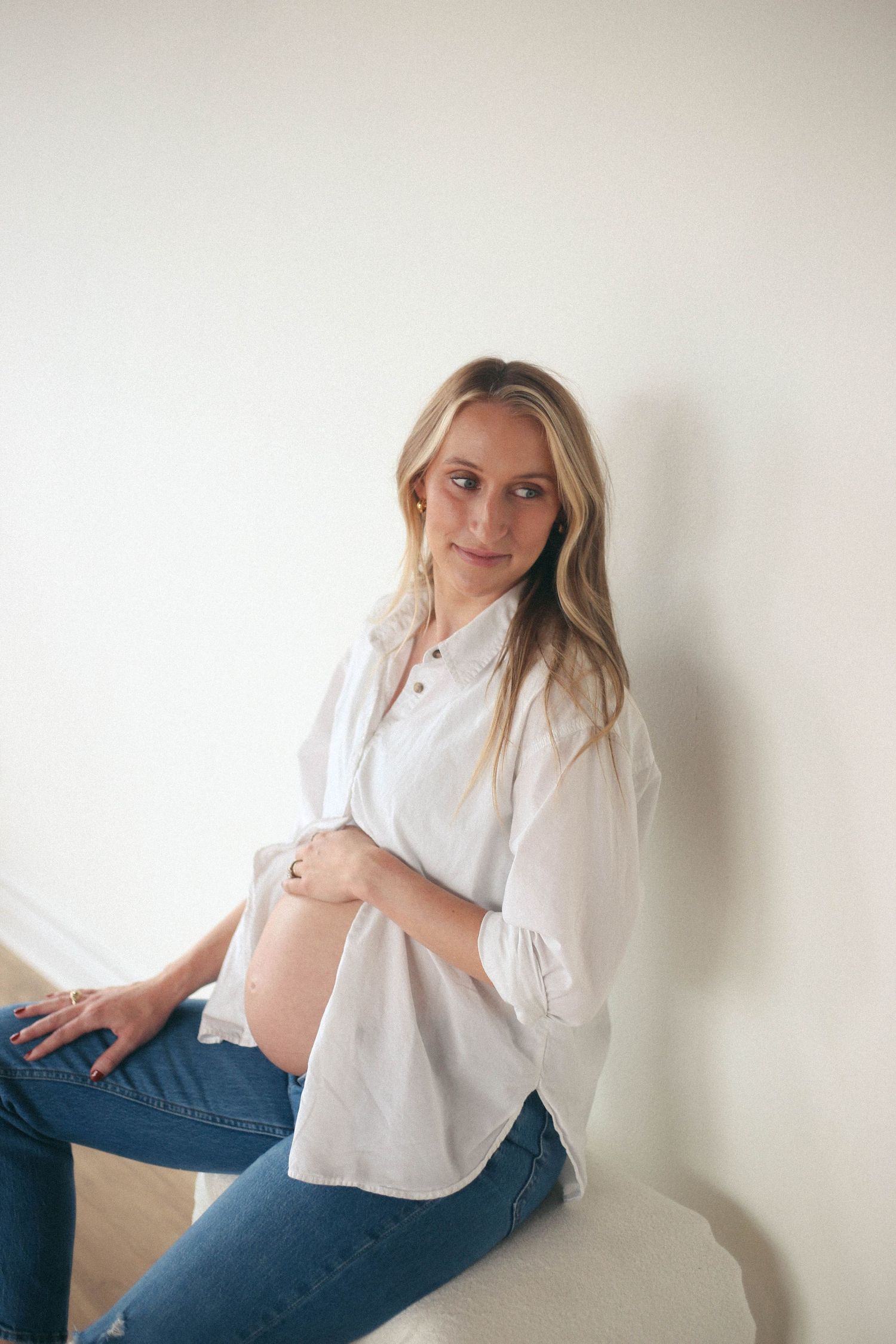 Pregnant woman in white blouse and jeans poses casually against a light wall in lifestyle portrait series.