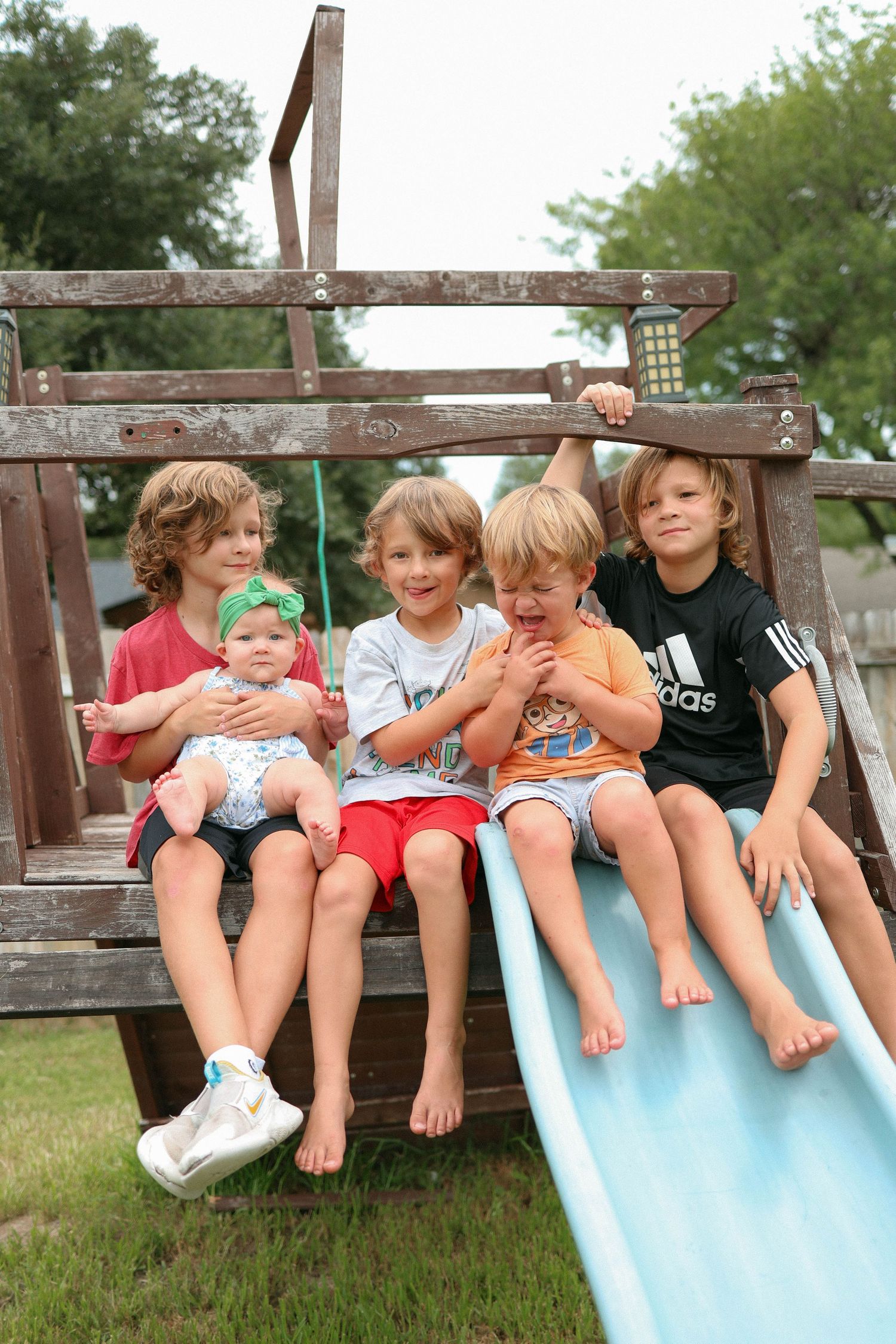 Five young children sit together on a wooden playground slide, smiling outdoors on a sunny day.