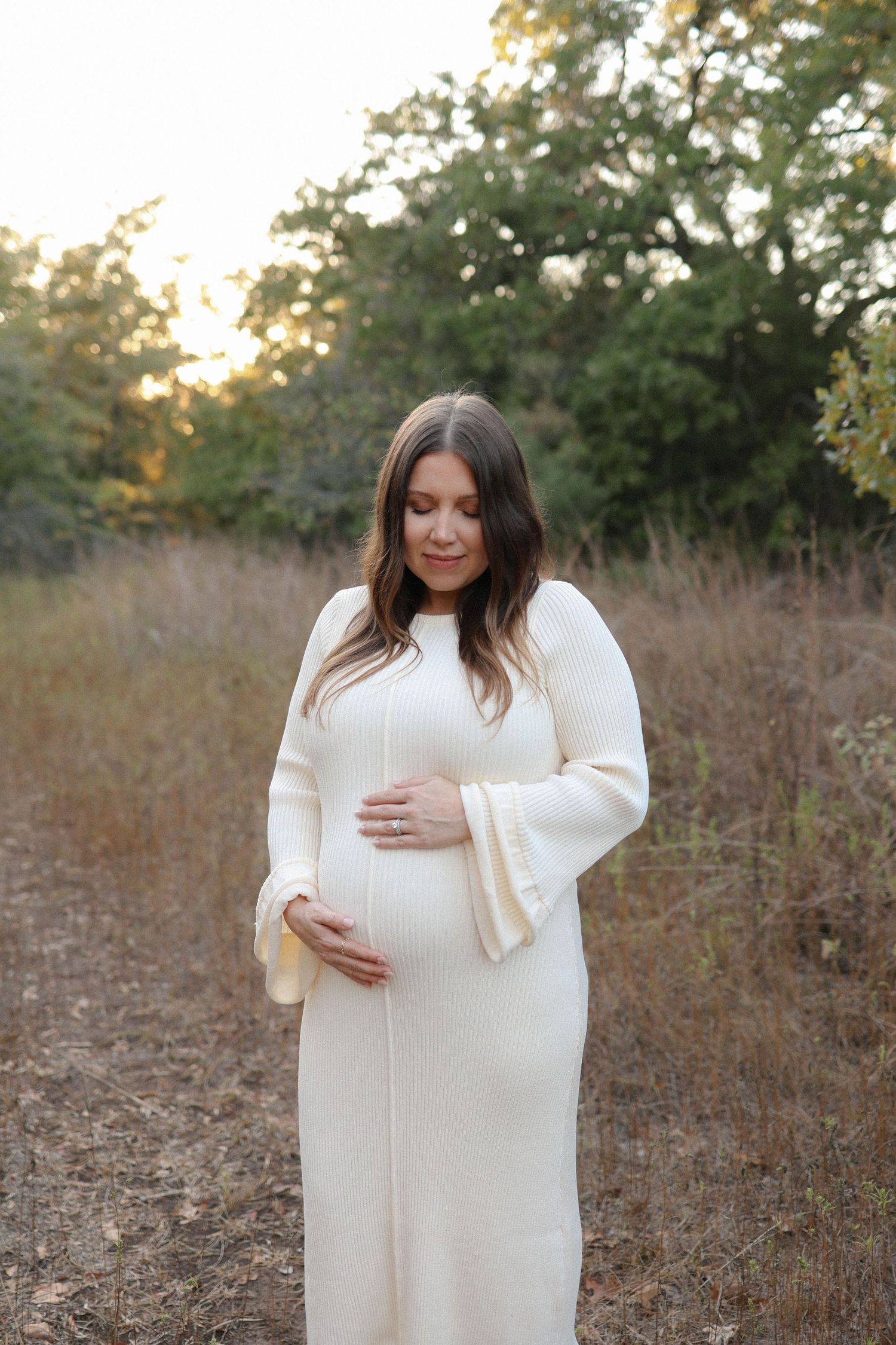 Pregnant woman in white maxi dress cradling baby bump outdoors in golden hour light among trees.