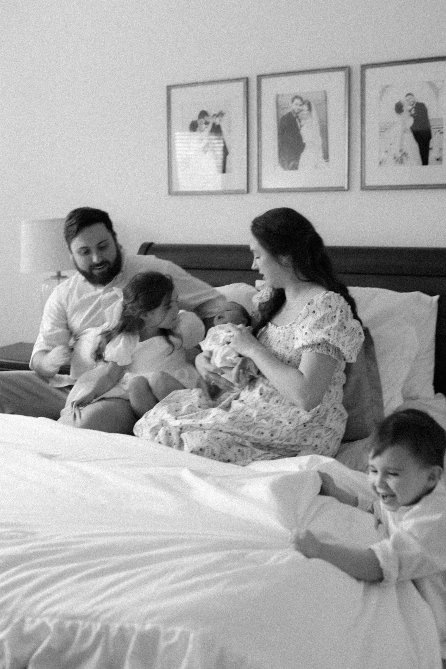 Black and white photo of a family with young children relaxing together on a bed with framed photos on the wall.