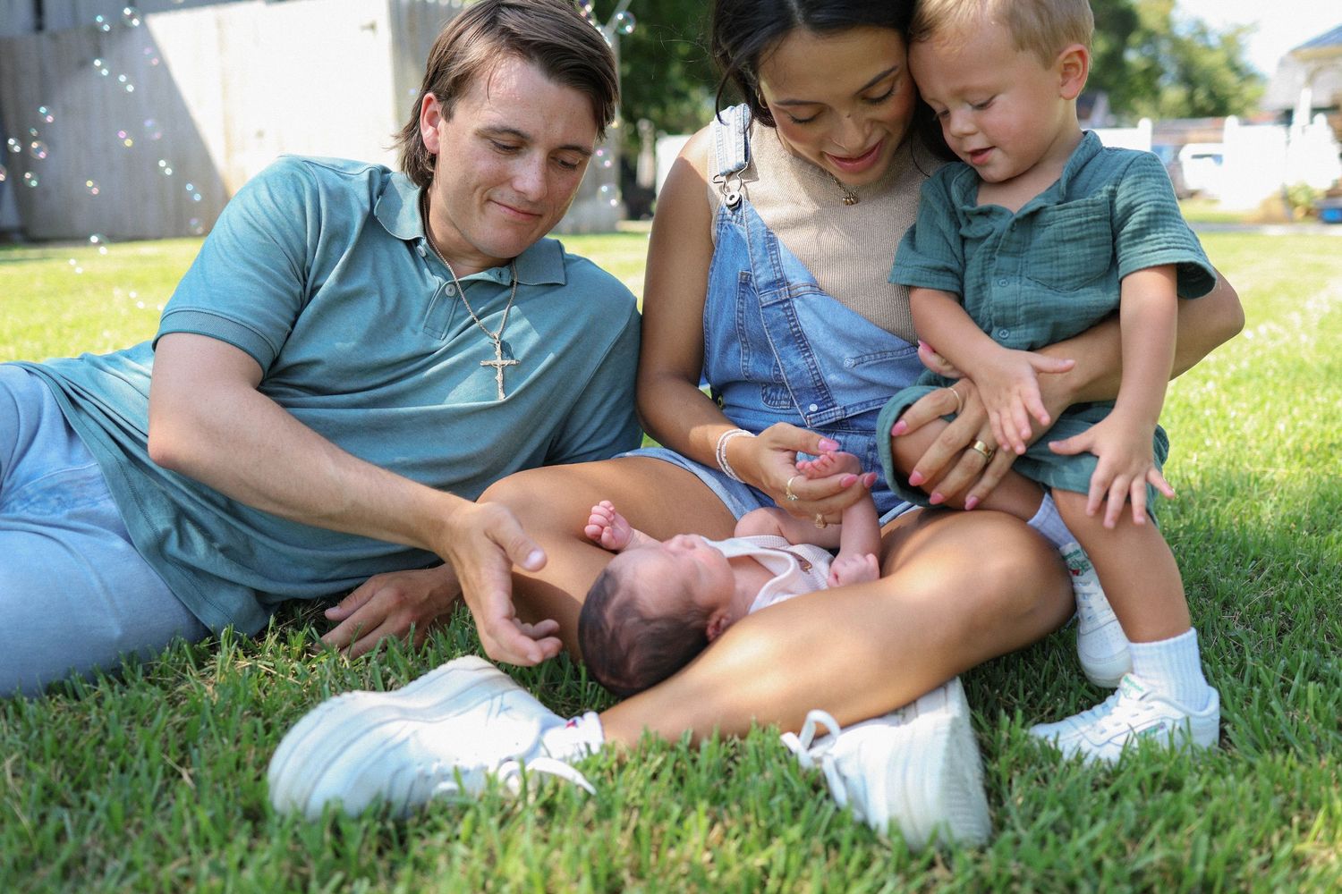 Family of four sitting on grass, parents smiling at newborn baby while toddler watches lovingly outdoors.