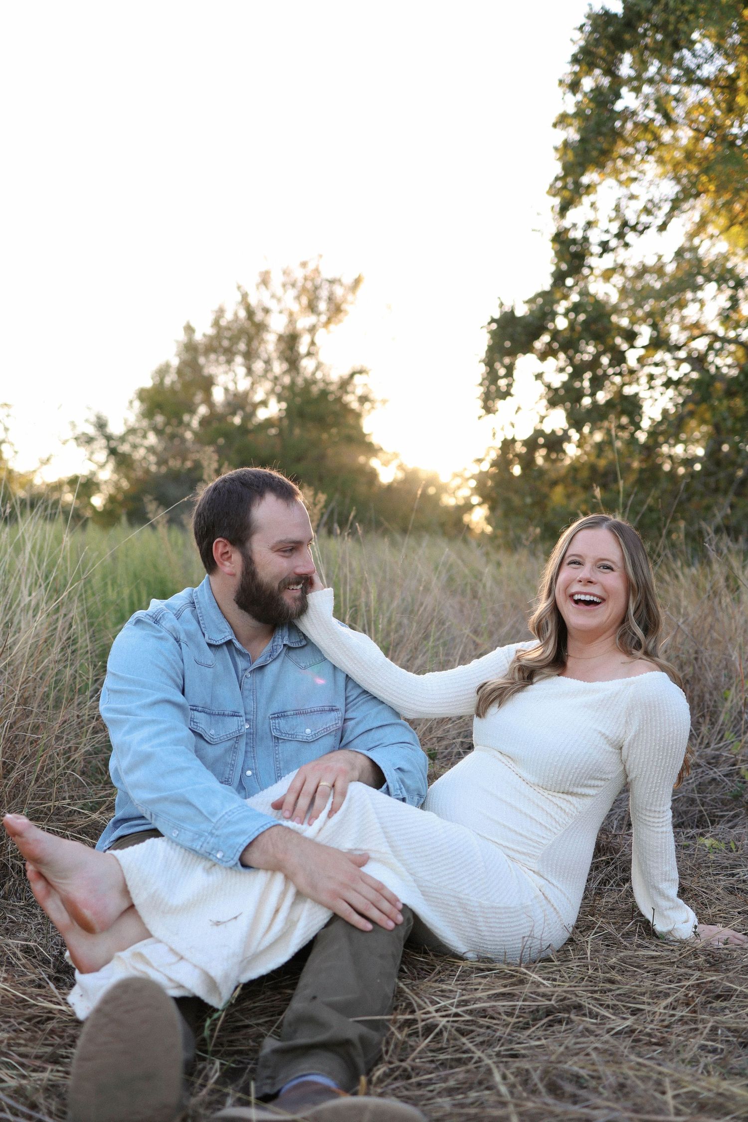 A laughing pregnant woman in a white dress sits with her partner in a golden grass field during a maternity photo session.