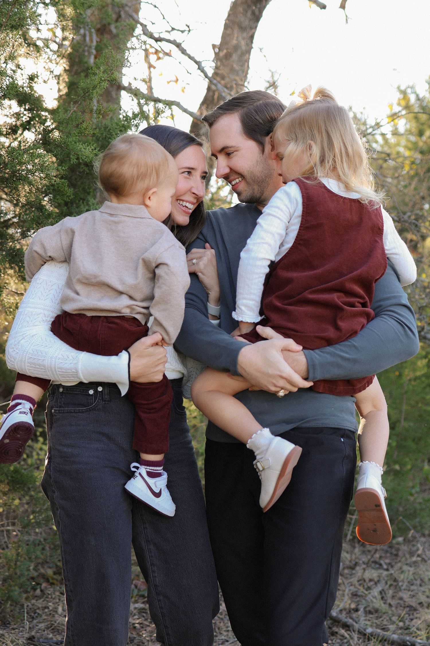 Happy family of four with two toddlers in matching burgundy vests sharing a joyful moment outdoors among trees.
