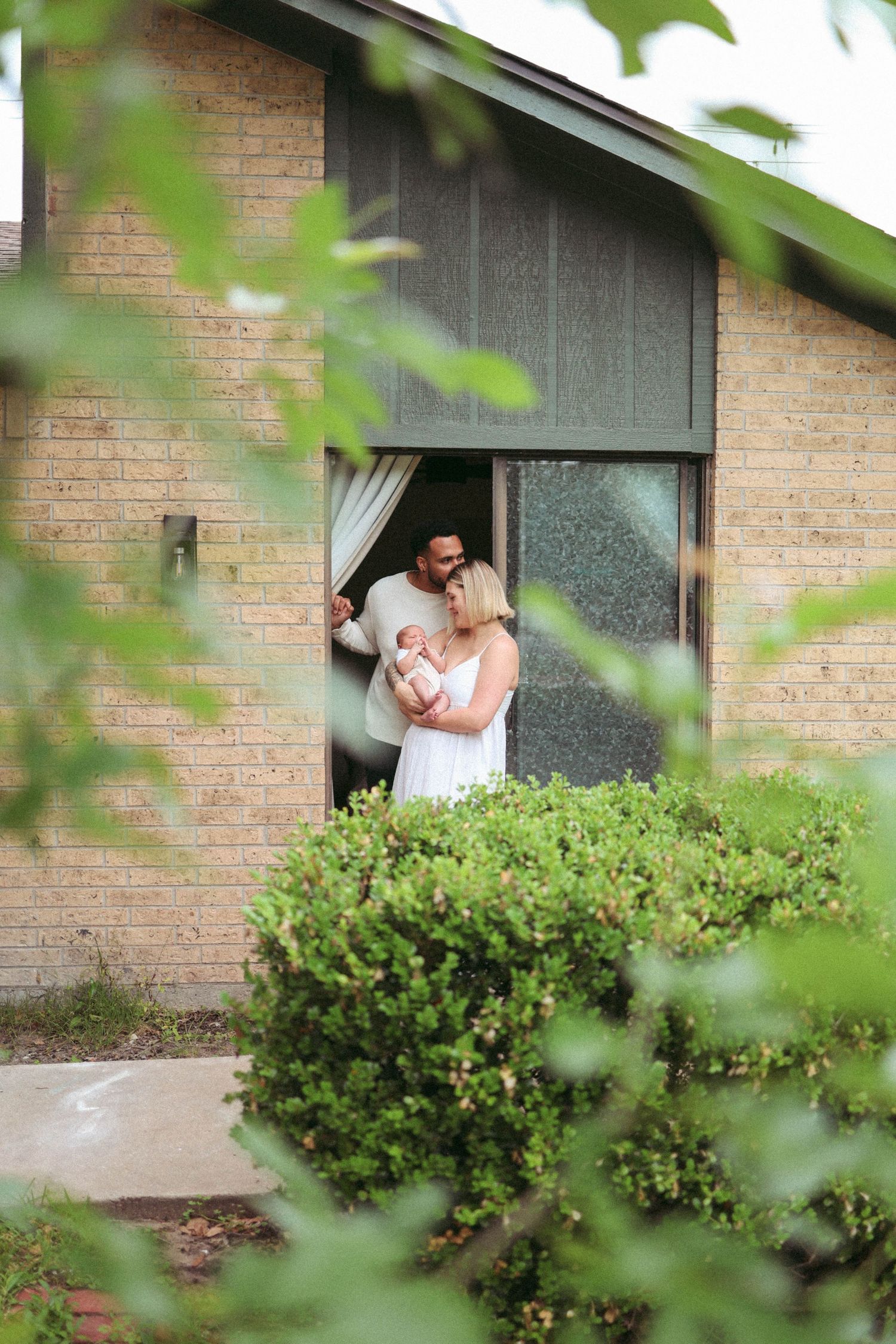 A family walking through greenery outside their brick home during a lifestyle newborn photography session.