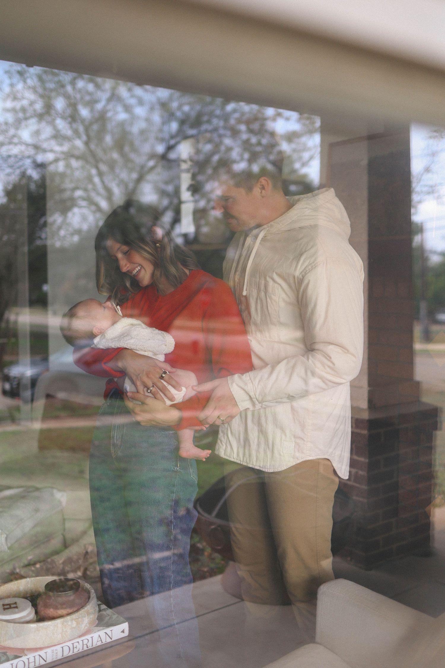 A couple smiles at their newborn baby, photographed through a window with reflections.