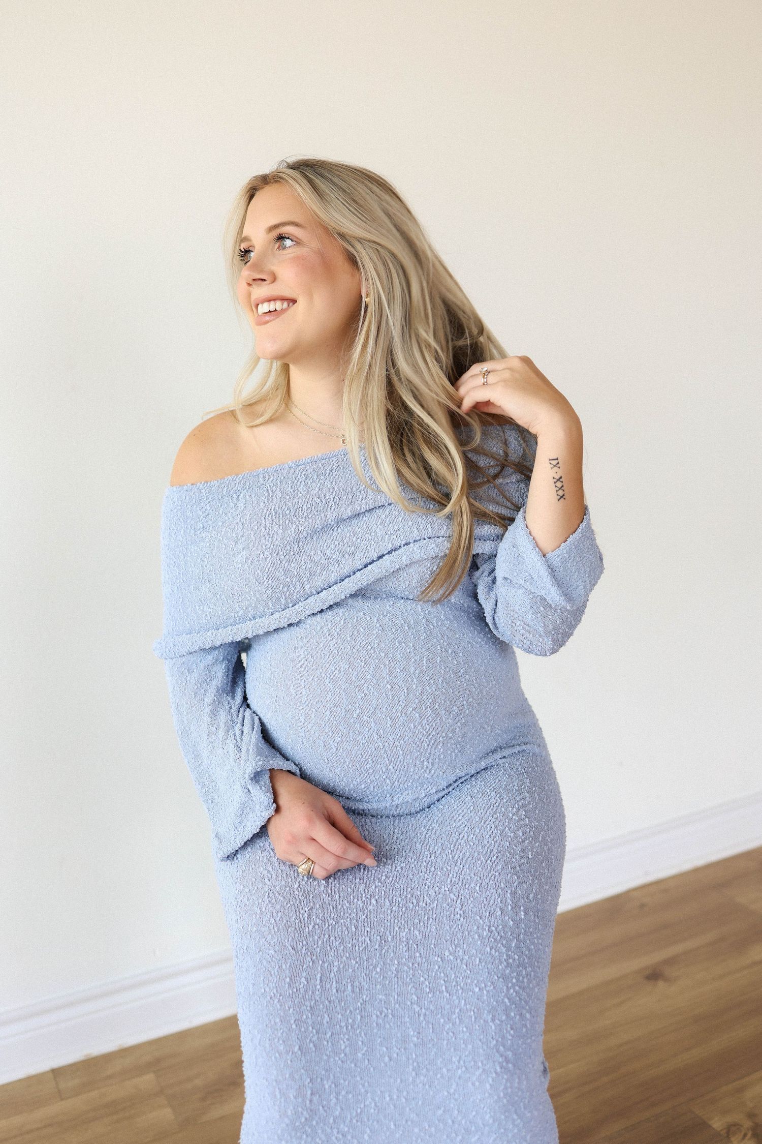 Smiling pregnant woman in a light blue off-shoulder textured dress, holding her baby bump, with long blonde hair.