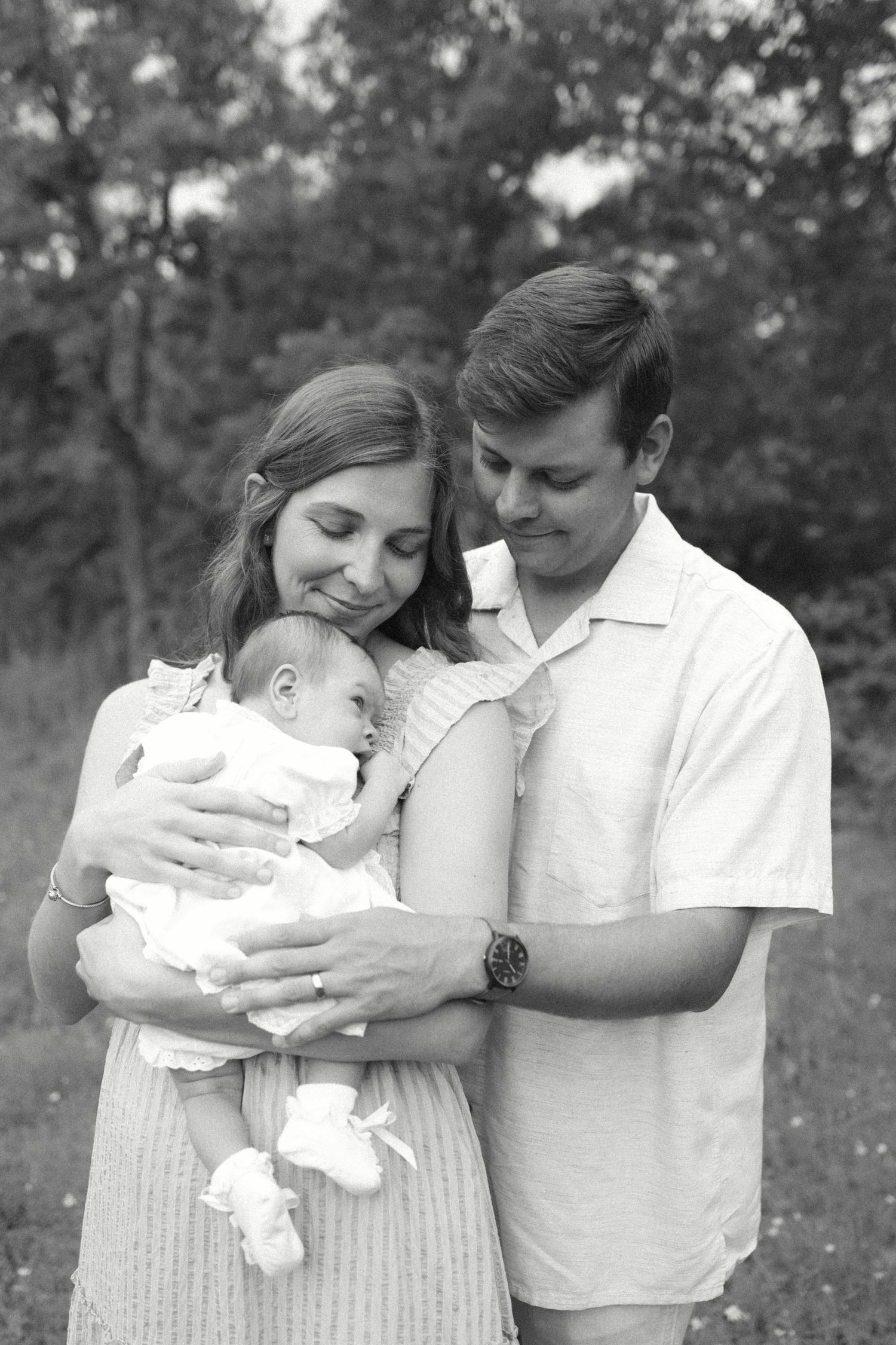 Black and white photo of young parents lovingly cradling their newborn baby outdoors in a lush green setting.