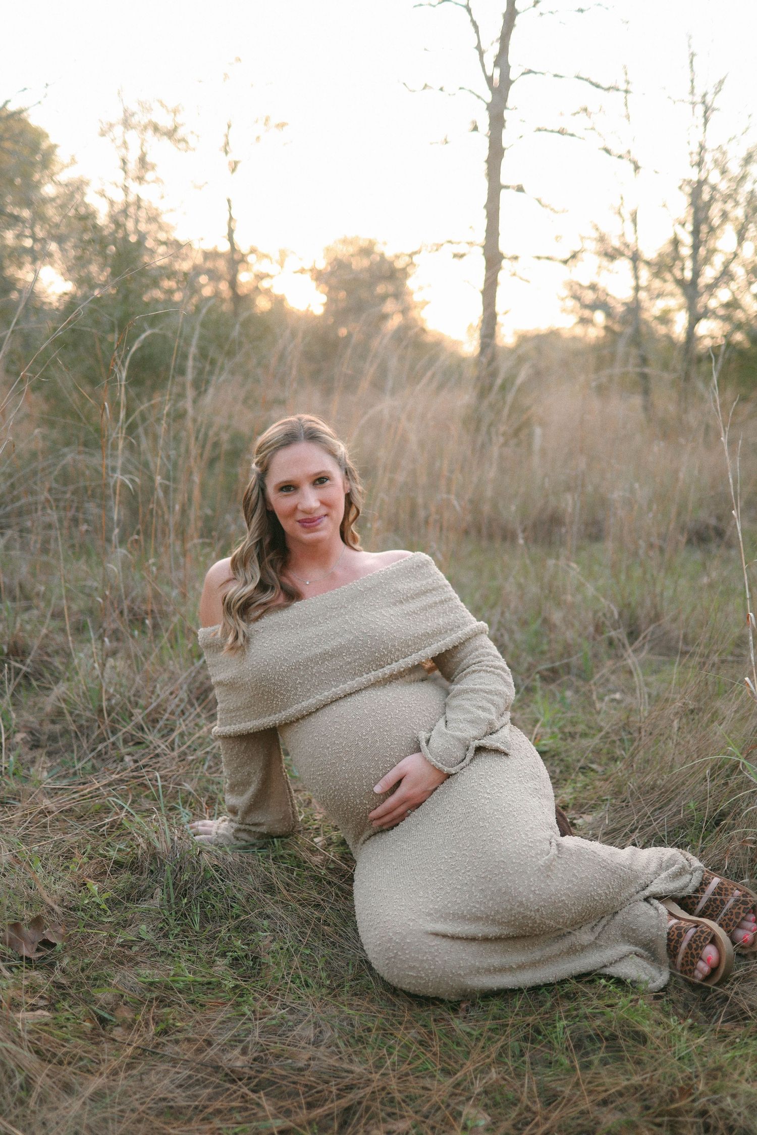 A pregnant woman in a beige sweater dress posing on the ground during an outdoor maternity photoshoot.