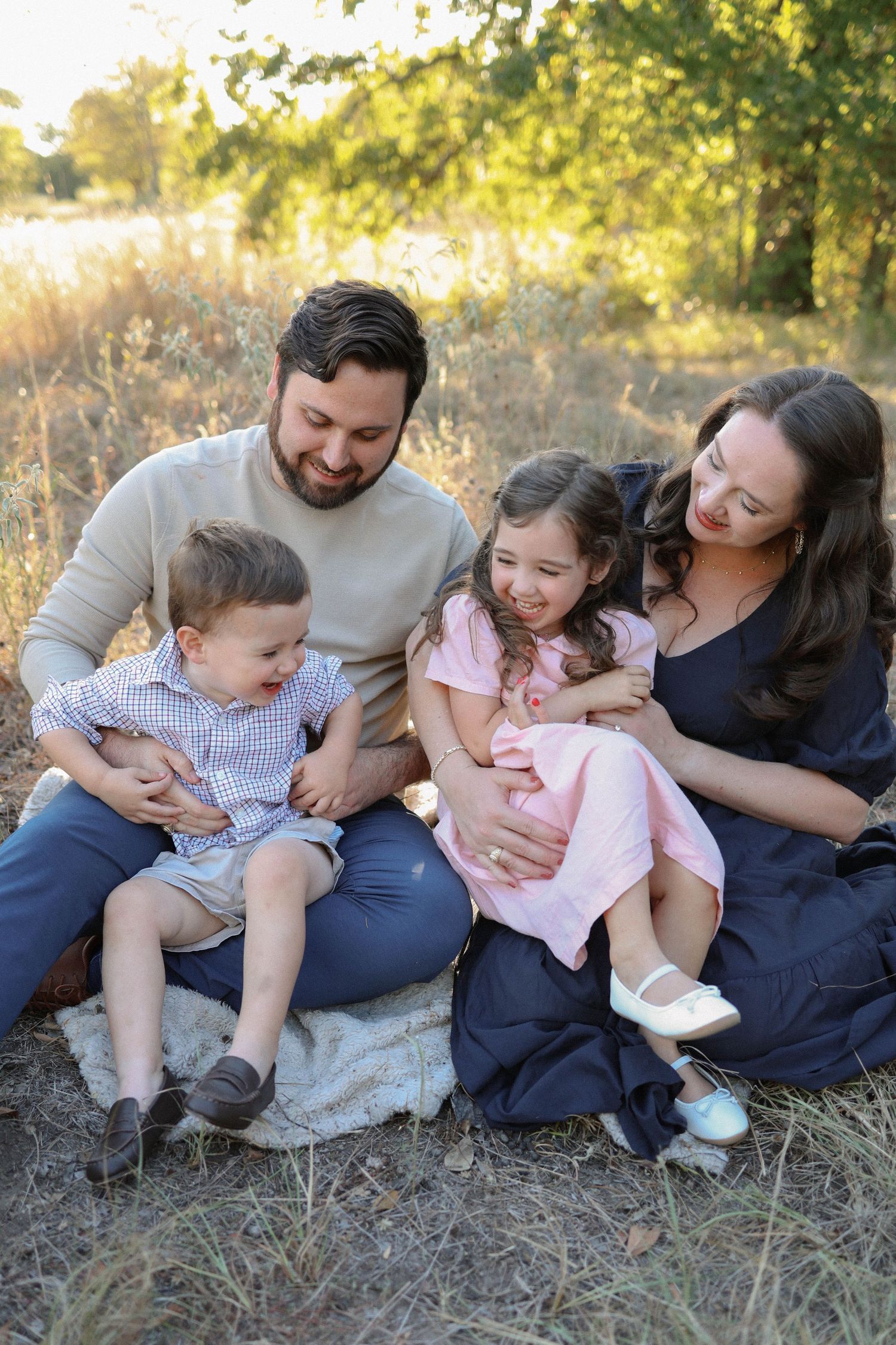 Happy family of four sitting outdoors in golden field, parents smiling with young son and daughter in pink dress.