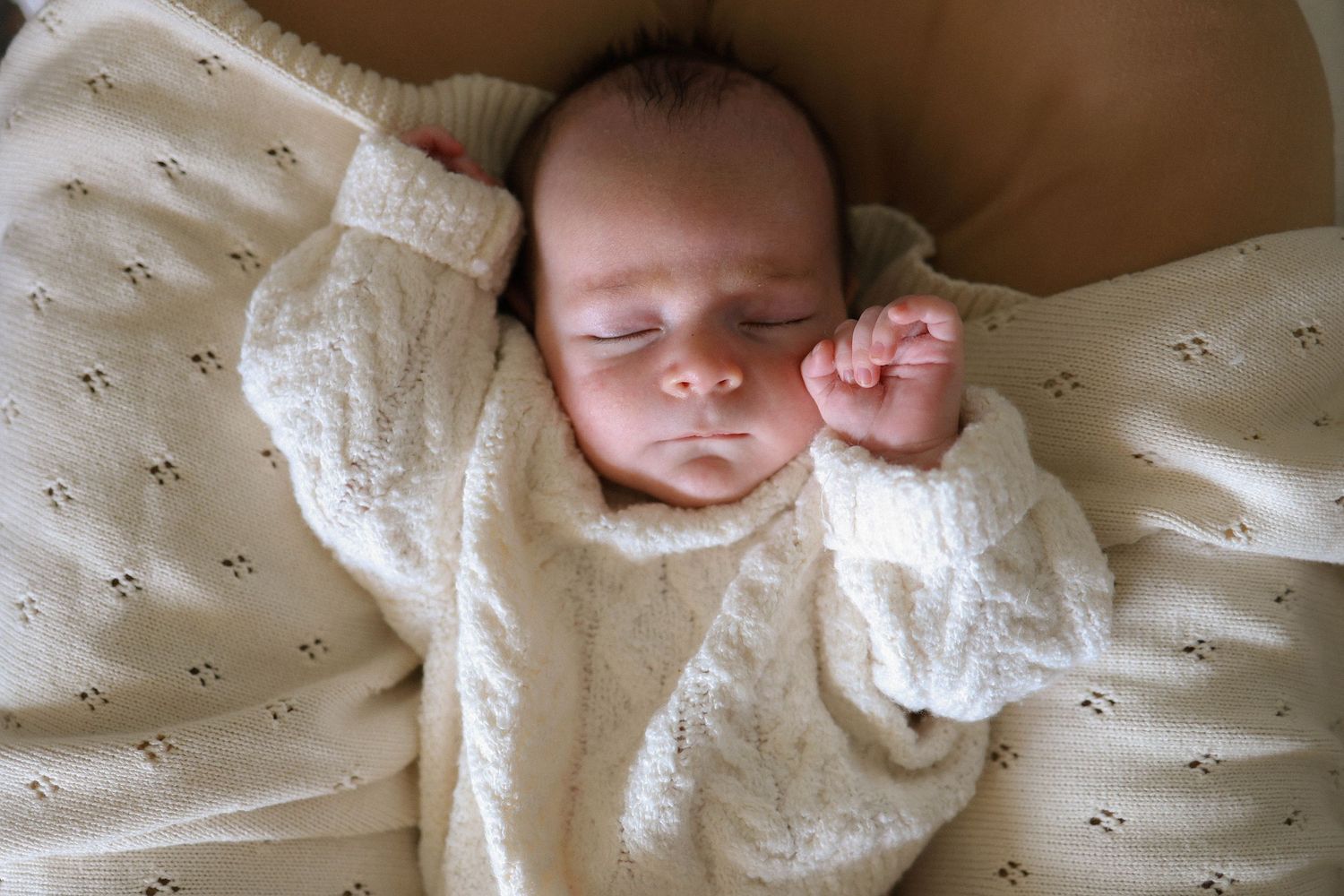 Peaceful newborn baby sleeping in a cream knit sweater on a soft white blanket.