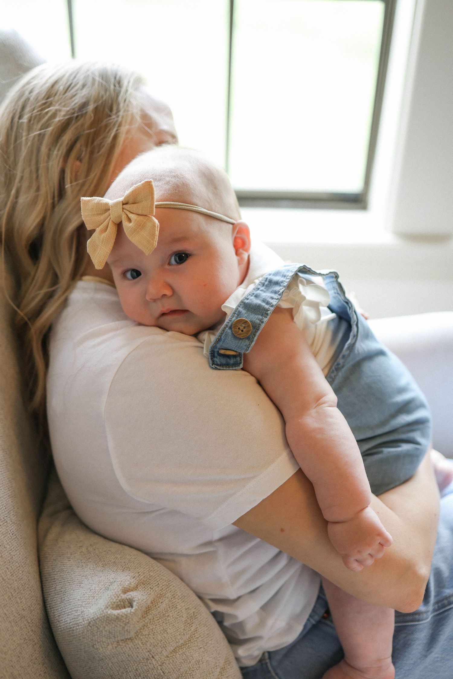 Baby cuddling against mom in white shirt by window.