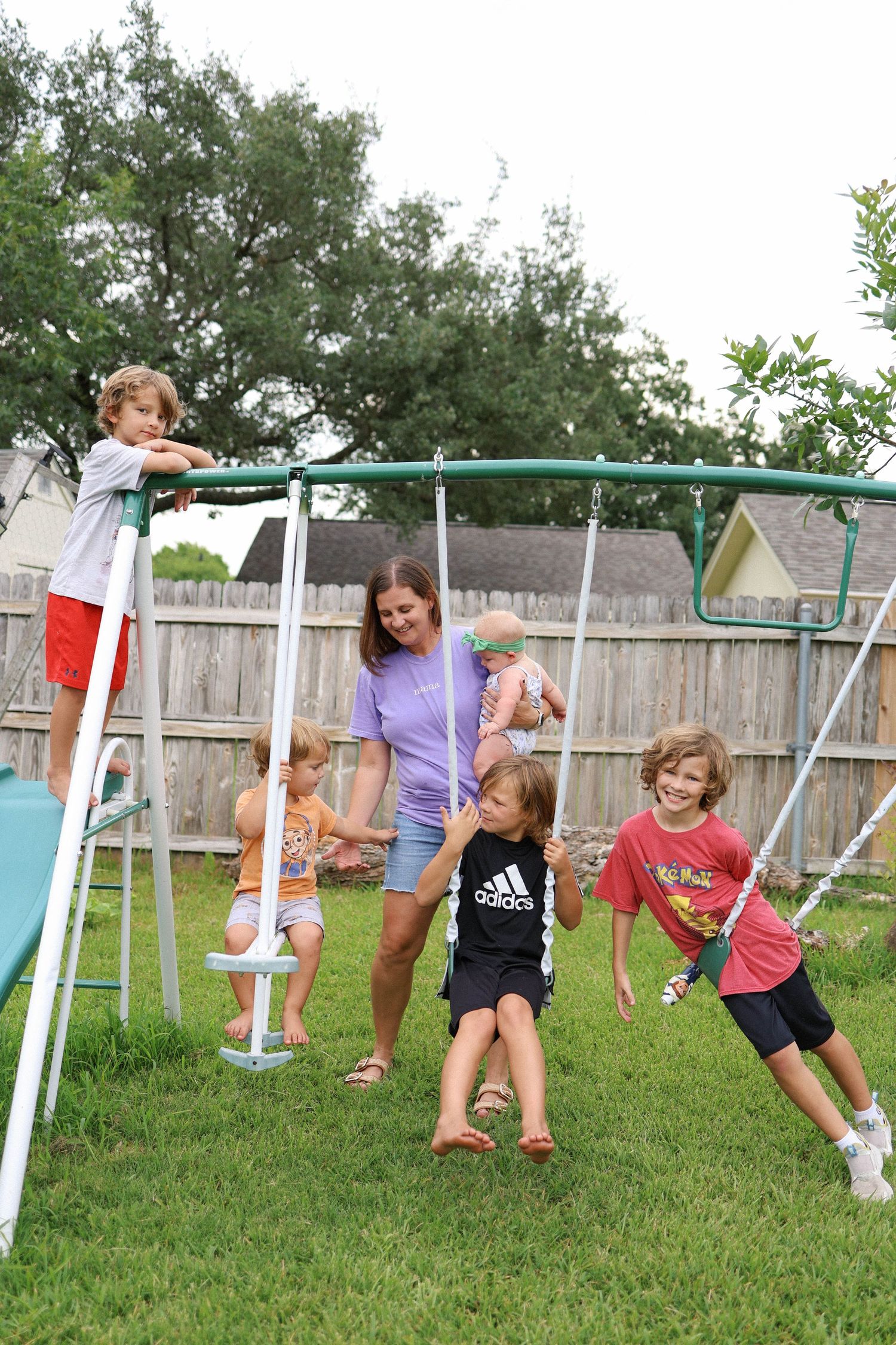 A woman supervises several children playing on a metal swing set in a fenced backyard on a cloudy day.