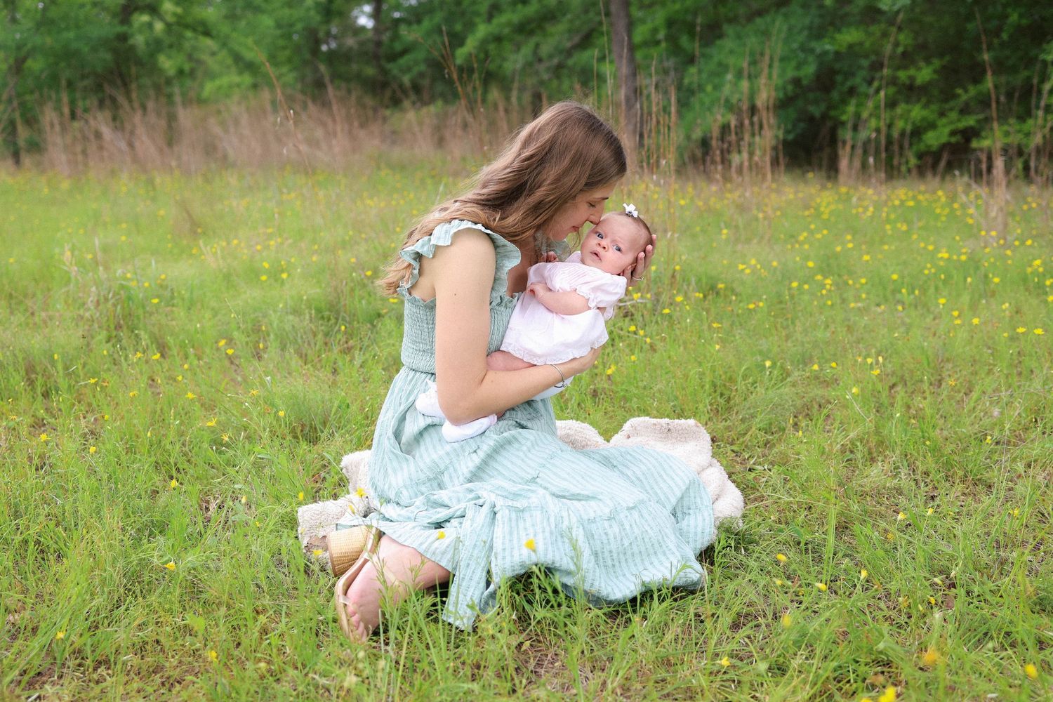 A young girl kneeling in a green meadow, tenderly kissing a newborn baby wrapped in a white blanket.
